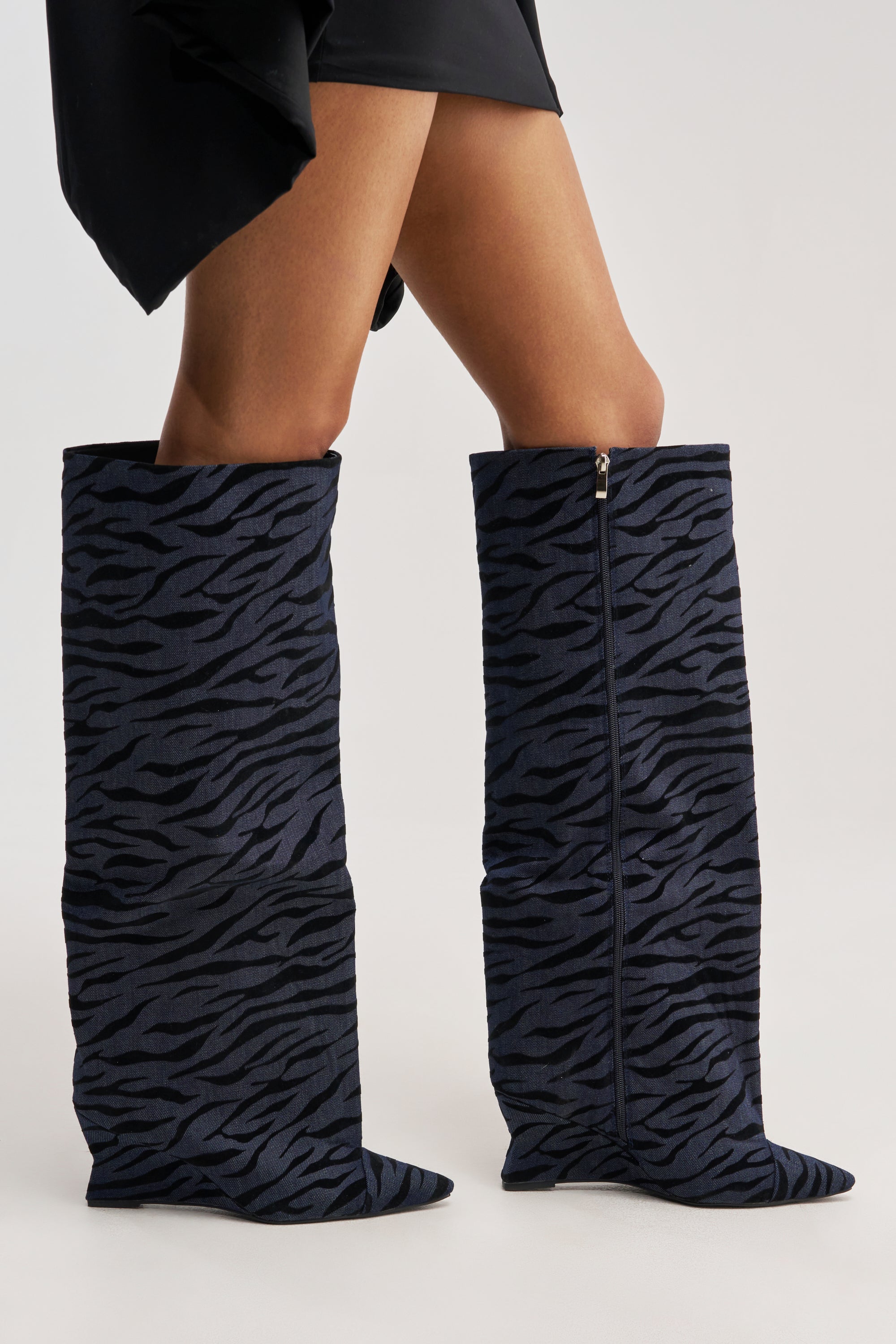 A person wears the AZALEA WANG KENYA DENIM TIGER WEDGE BOOT, featuring a tiger print with dark blue and black zebra patterns, styled with a black skirt and posed against a plain light background.
