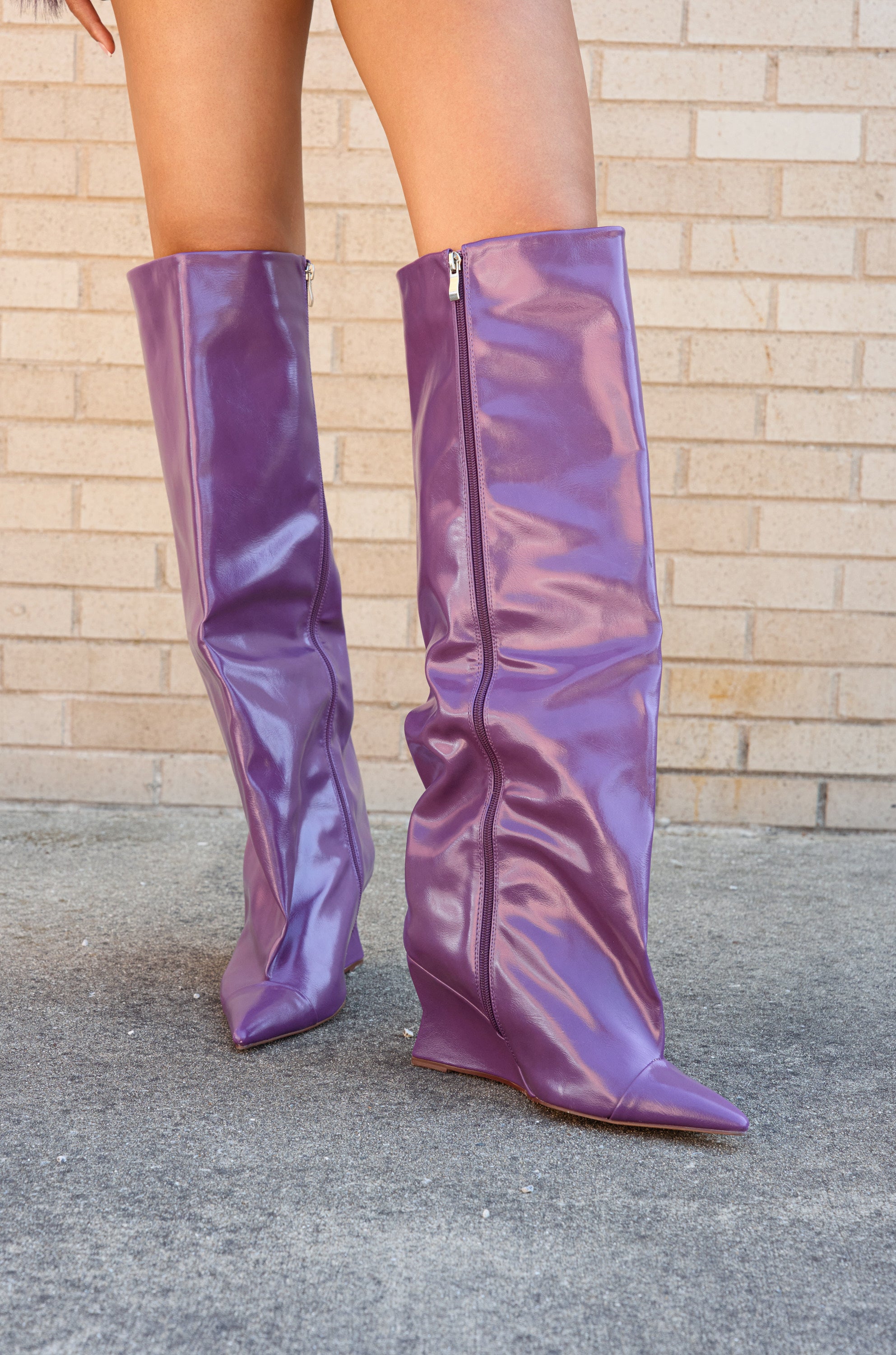 A person stands on concrete in front of a light brick wall, wearing the AZALEA WANG KENYA PURPLE KNEE HIGH WEDGE BOOT with a shiny finish and pointed toes.