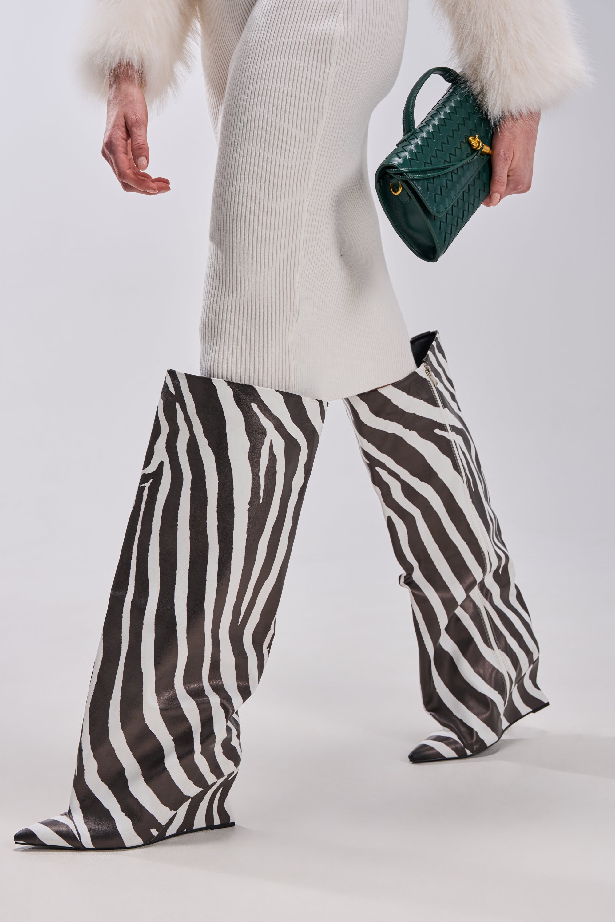 A person in a white ribbed skirt, white fur-cuffed sleeves, AZALEA WANG KENYA ZEBRA KNEE HIGH WEDGE BOOTs, and holding a textured green handbag walks on a plain white background.