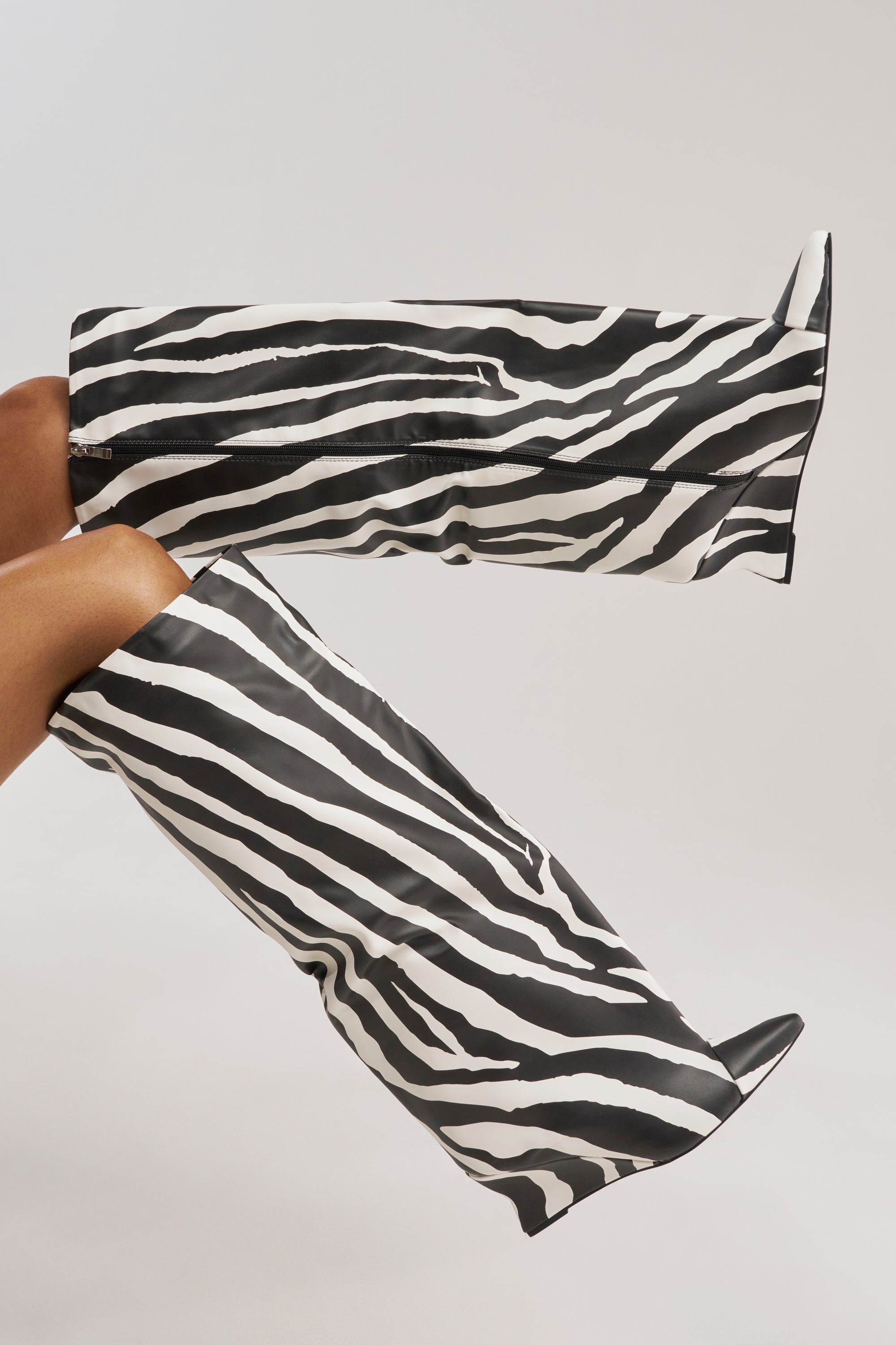 A person wears the AZALEA WANG KENYA ZEBRA KNEE HIGH WEDGE BOOT, featuring knee-high, pointed toes and angular structure in bold black and white faux leather zebra print, set against a plain light background.