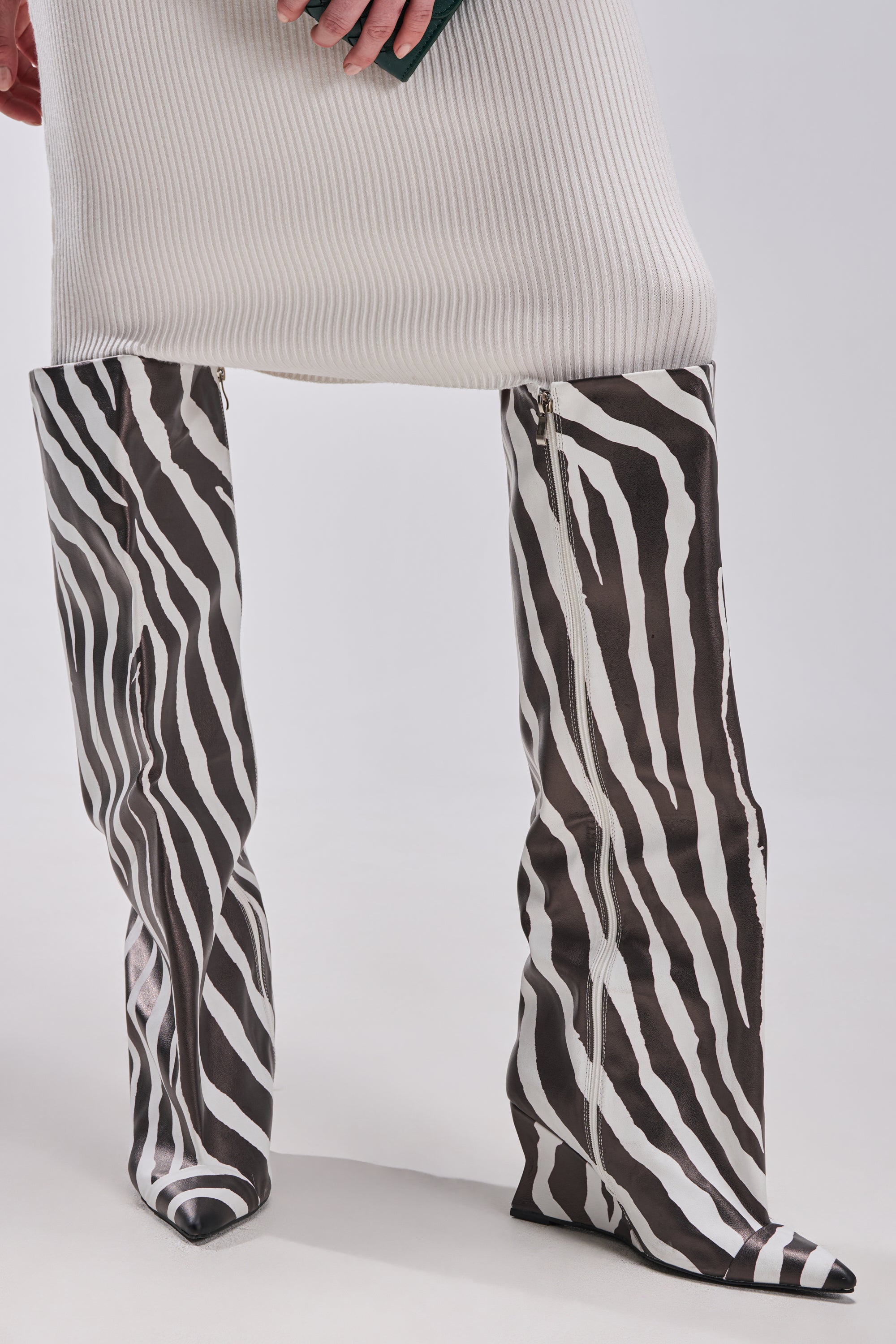 A person in a ribbed white skirt and AZALEA WANG KENYA ZEBRA KNEE HIGH WEDGE BOOT with pointed toes stands against a plain light background.