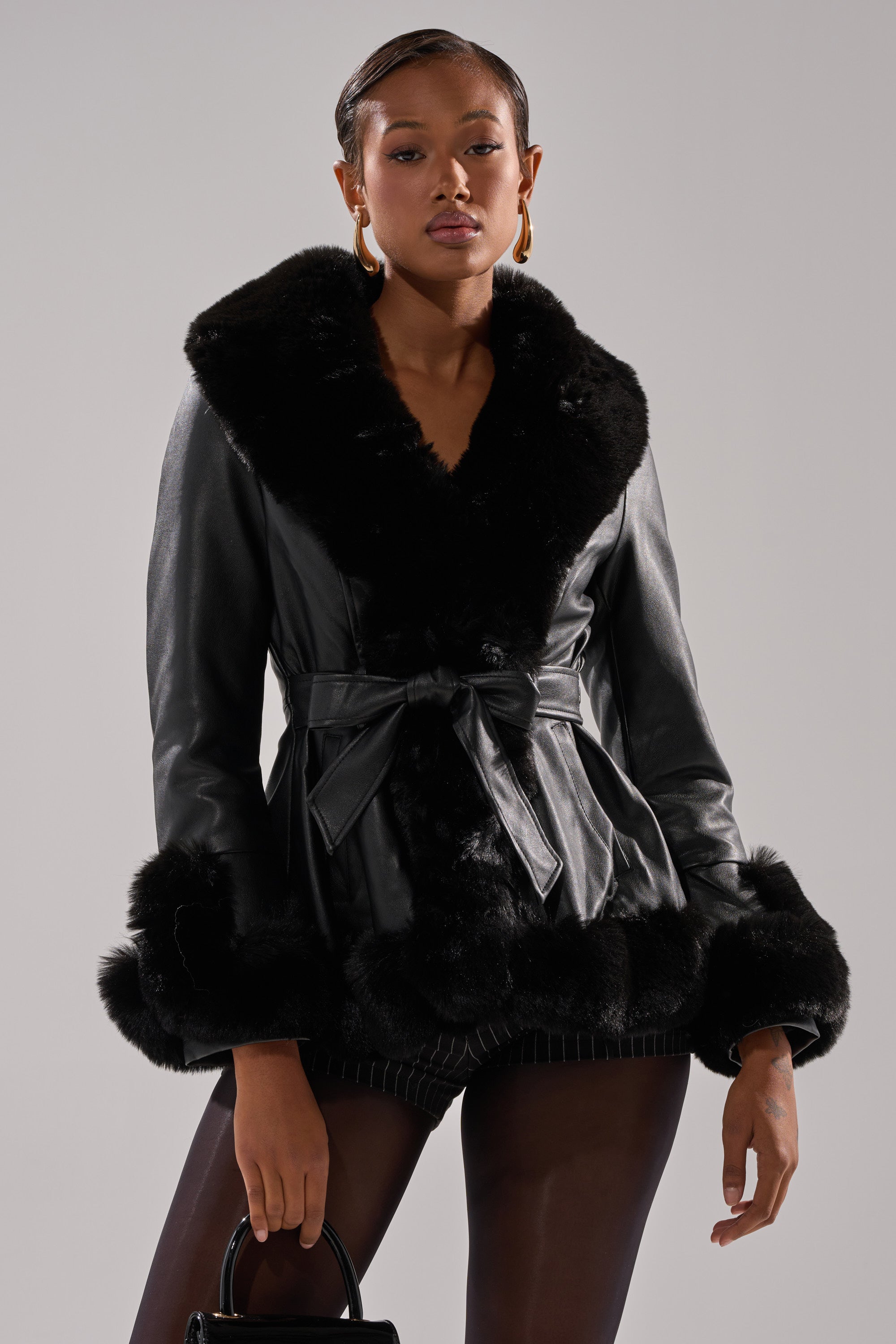 Wearing the KENZY FUR TRIM PEPLUM MOTO IN BLACK, belted at the waist with faux fur trim, a person styles it with dark tights, gold hoop earrings, and a black handbag against a neutral background.