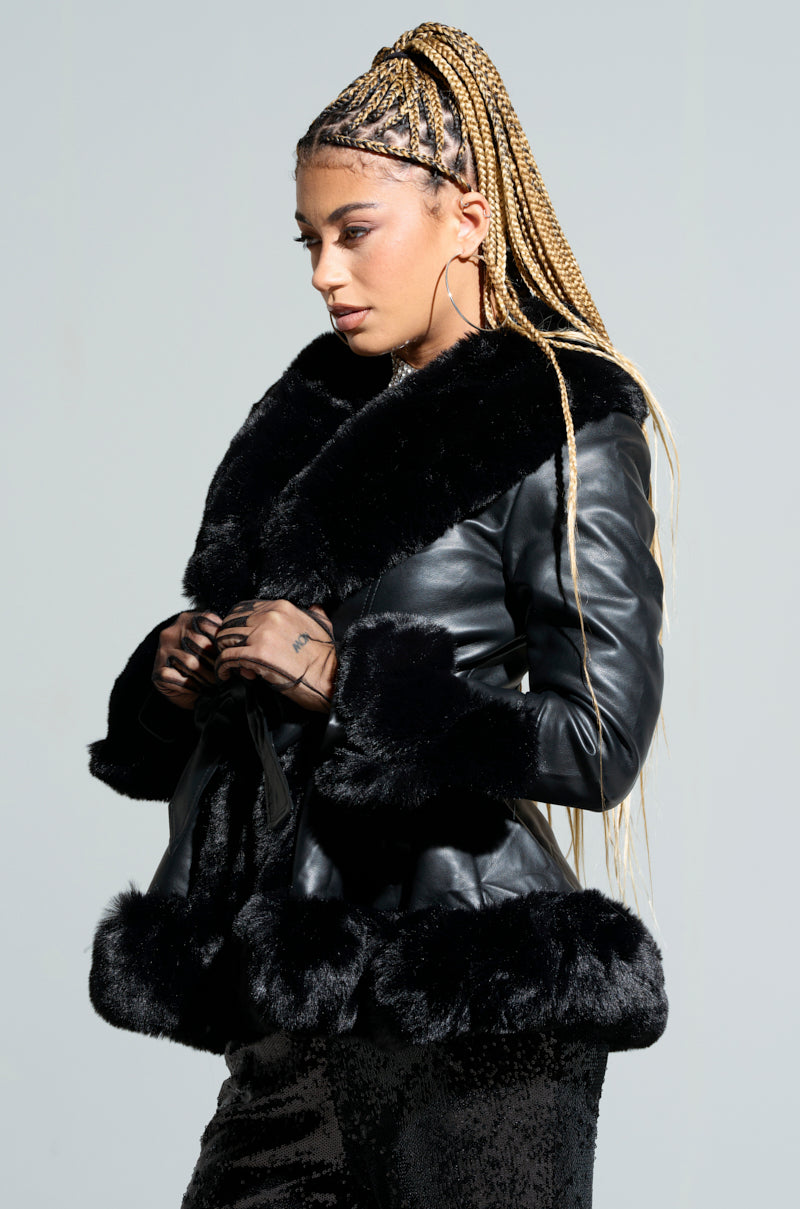 A person with long braided hair wearing the KENZY FUR TRIM PEPLUM MOTO IN BLACK and dark sequin pants poses against a neutral background. They appear contemplative, with hands gently holding the front of the peplum moto jacket.