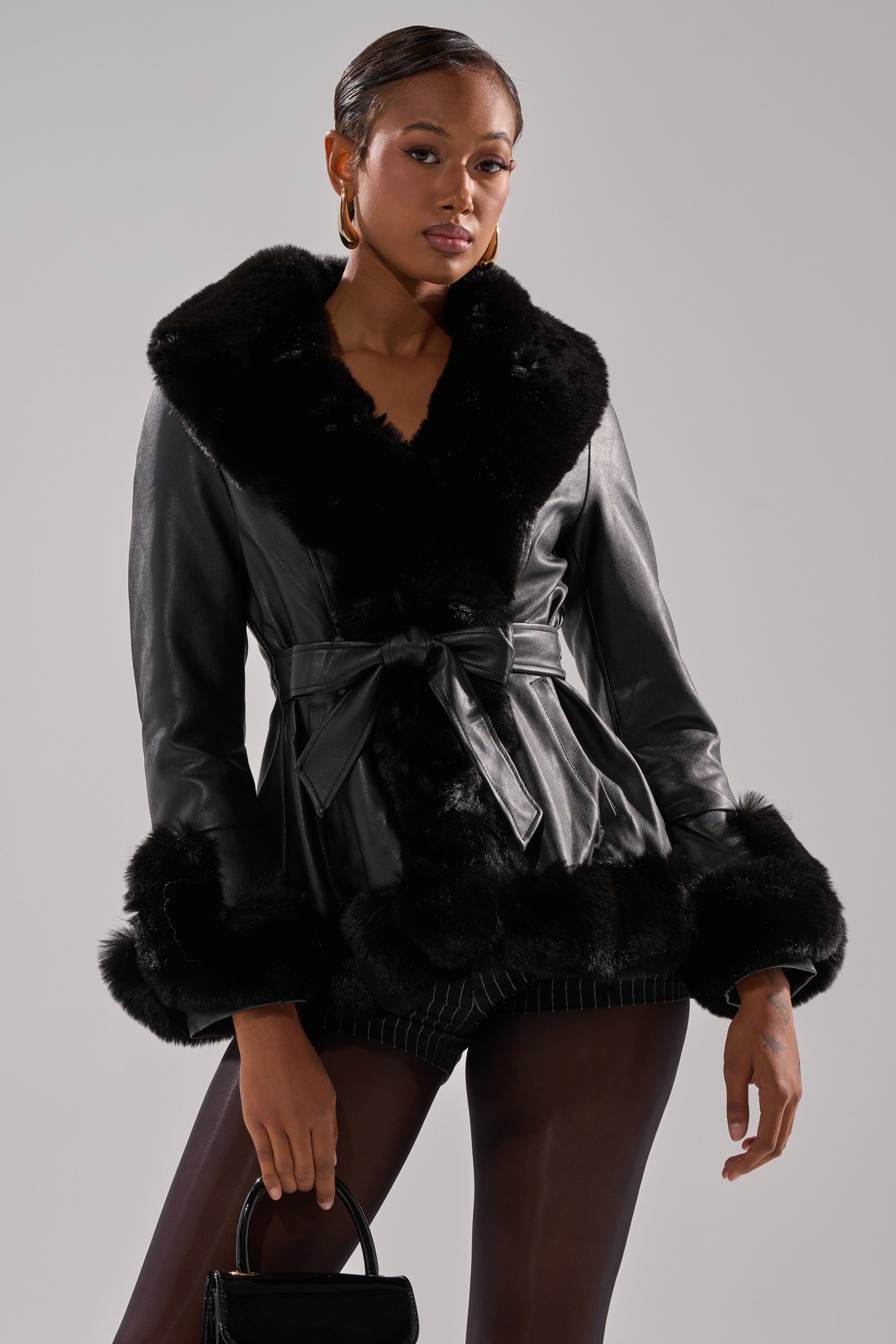 Wearing the KENZY FUR TRIM PEPLUM MOTO IN BLACK, a woman poses against a light gray background. The jacket features a large faux fur collar and cuffs, a waist tie belt, and she accessorizes with a small black handbag.