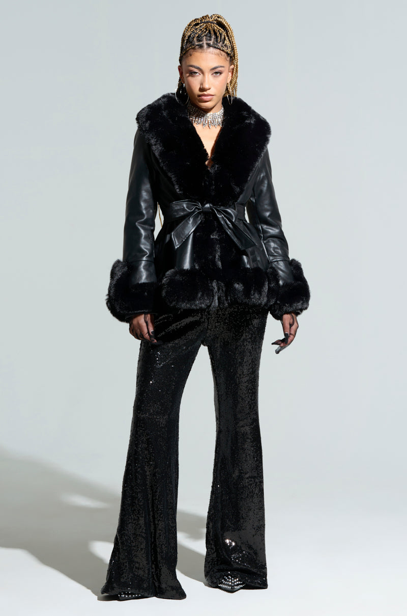 Wearing the Kenzy Fur Trim Peplum Moto in Black, paired with sequined pants, a person exudes confidence. Their braided hair is highlighted by a choker necklace against a light gray background. The ensemble skillfully combines the textures of faux fur and sleek leather jacket accents.