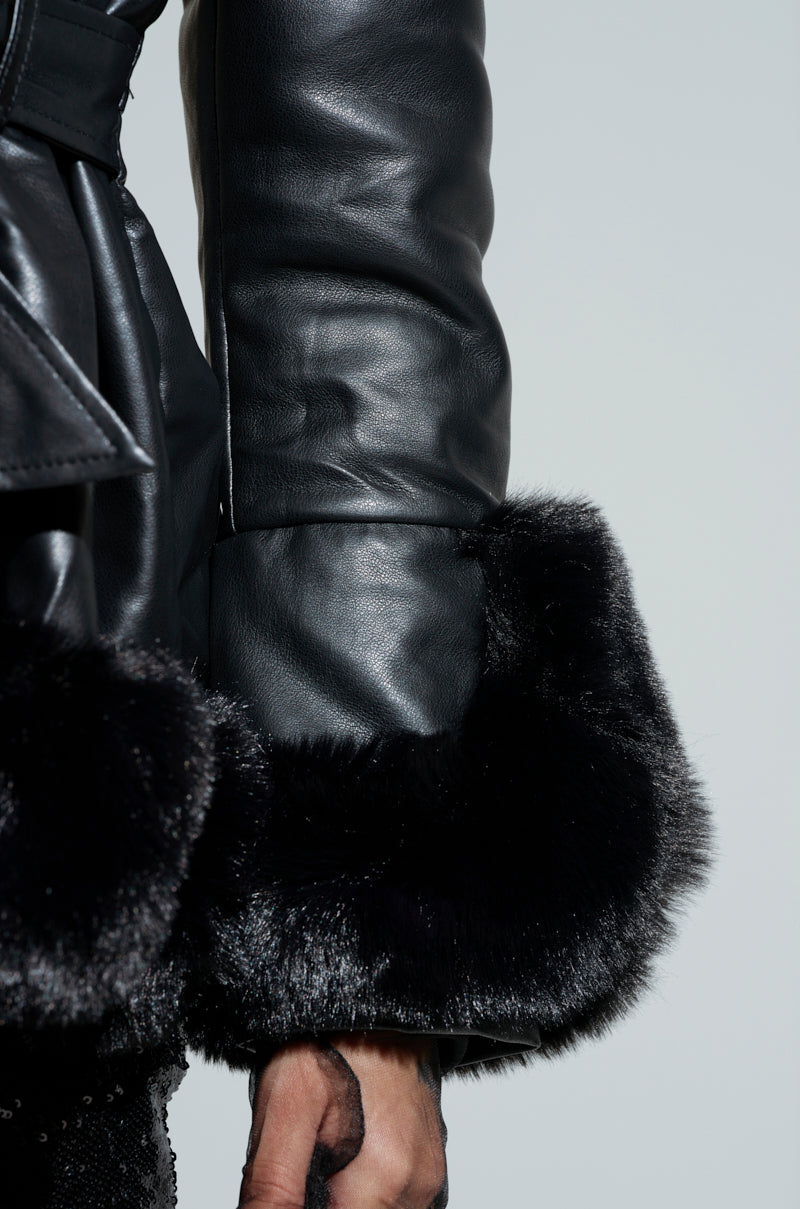 A close-up shows someone wearing the KENZY Fur Trim Peplum Moto in Black, featuring glossy black leather with plush faux fur trim on the sleeves and hem. Their hand rests casually at their side, highlighting the peplum moto design against a plain, light-colored background.