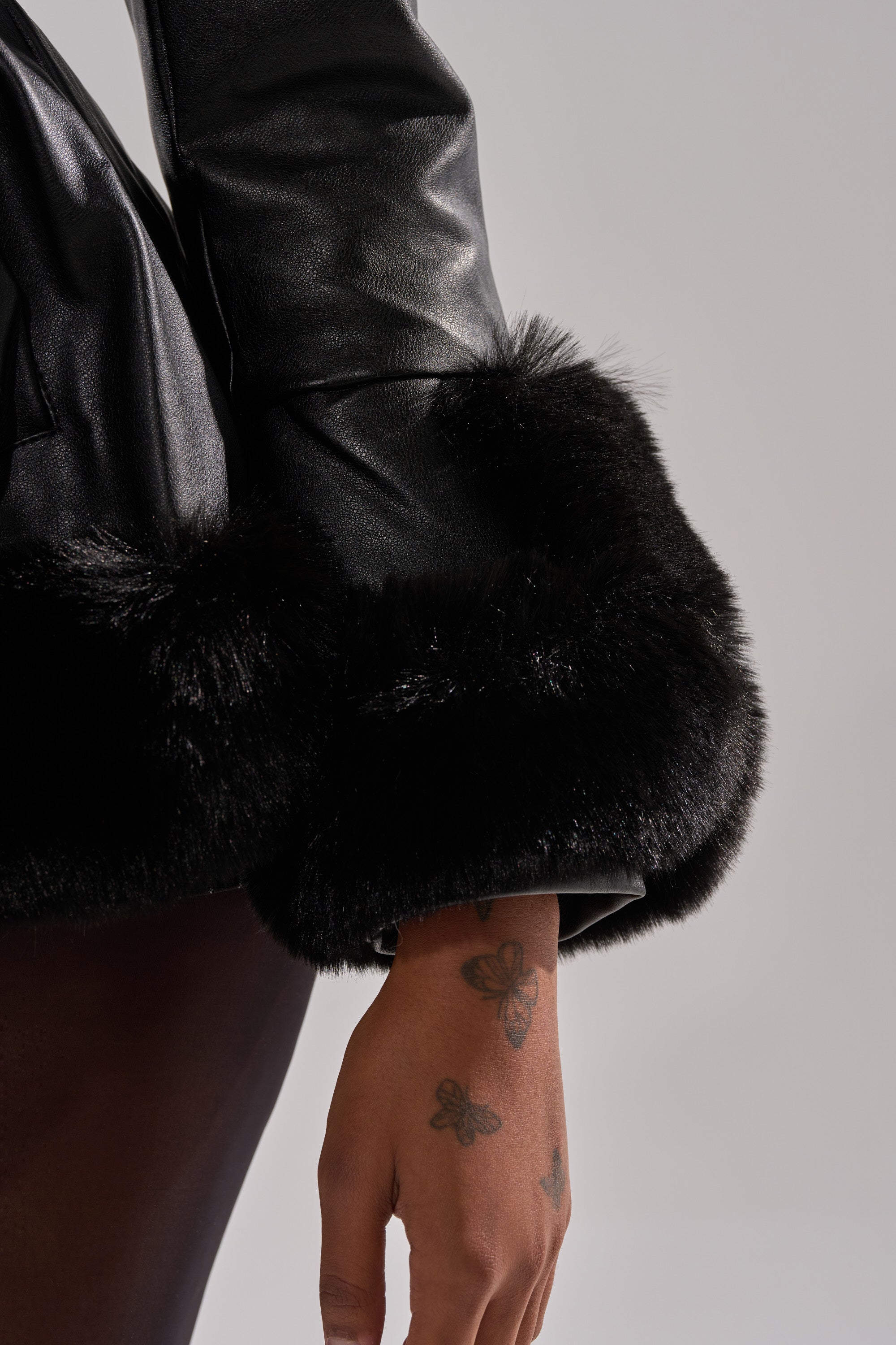 A close-up shows a person’s arm in the KENZY FUR TRIM PEPLUM MOTO IN BLACK, featuring black fur-trimmed sleeves. Their hand with butterfly tattoos is visible, highlighting this chic jacket against a plain background.