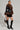 A person stands confidently, adorned in the KENZY FUR TRIM PEPLUM MOTO IN BLACK BROWN, paired with sleek black knee-high boots. They carry a stylish brown handbag and have their hair pulled back, set against a neutral background.