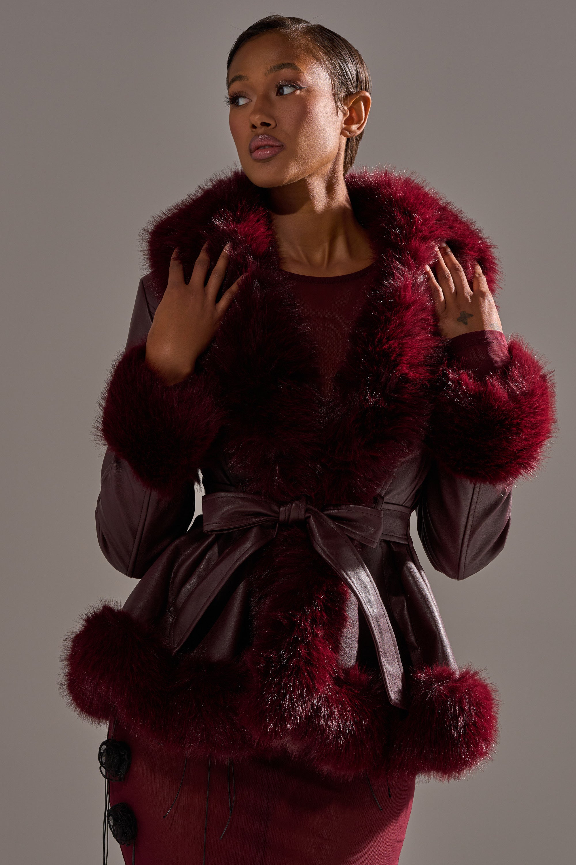 A woman poses against a gray backdrop, wearing the KENZY FUR TRIM MOTO IN BURGUNDY with a plush fur collar and cuffs, tied at the waist. She looks to the side with one hand near her collar. 2