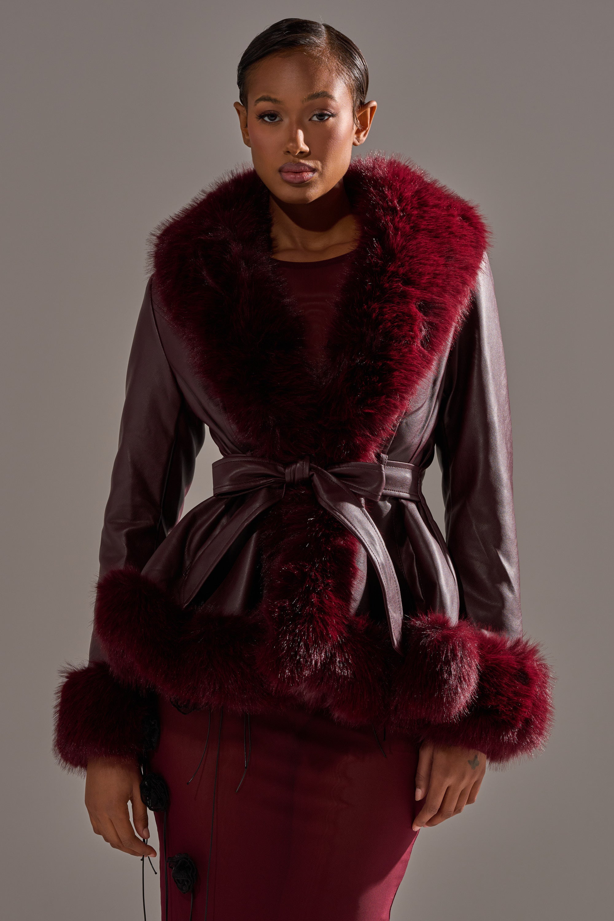 A woman wears the KENZY FUR TRIM MOTO IN BURGUNDY with a matching belt and dress, standing against a plain gray background. 0