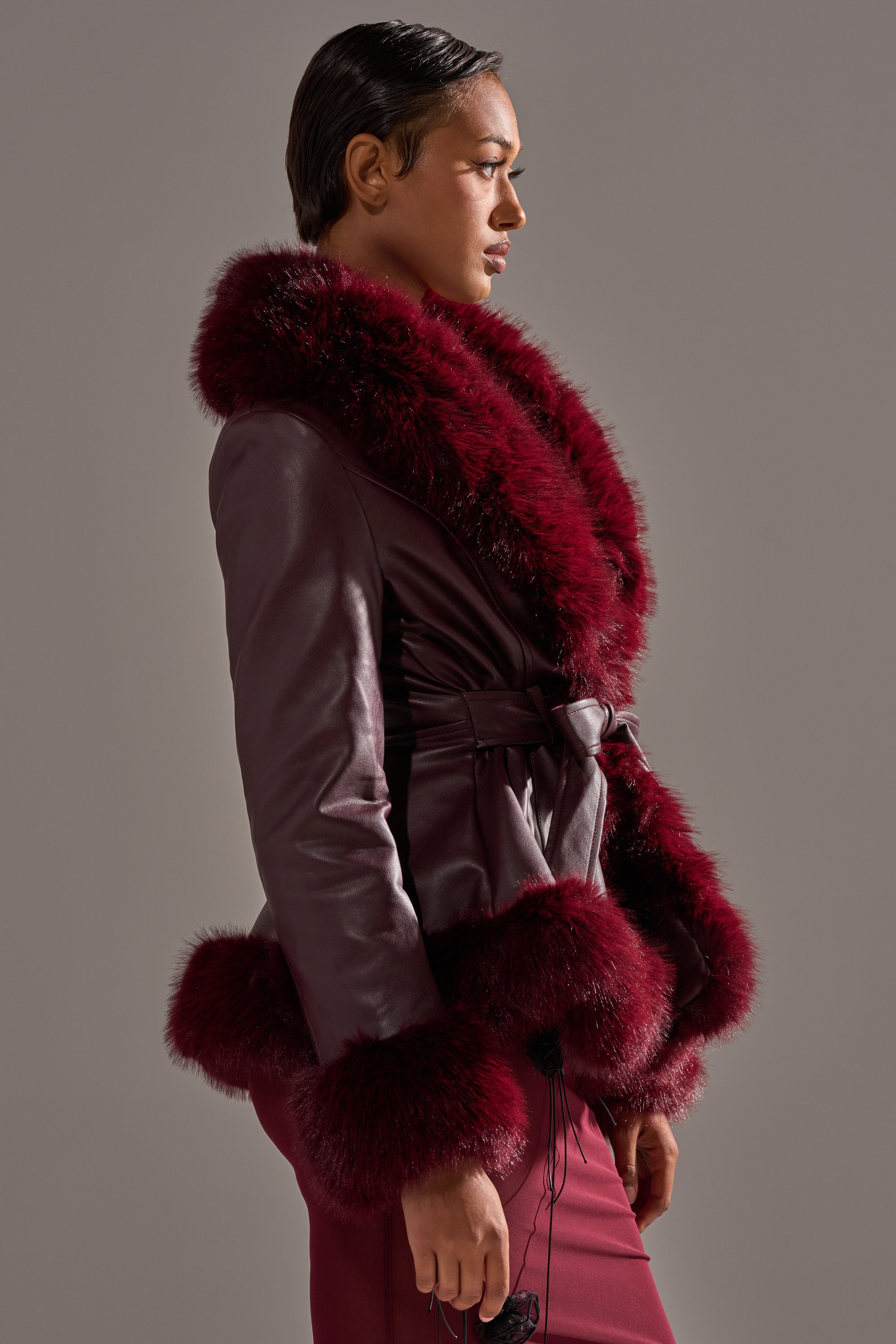 A person in profile models the KENZY FUR TRIM MOTO IN BURGUNDY—featuring plush fur trim on the collar, cuffs, and hem—with a matching skirt, set against a plain gray background. 4