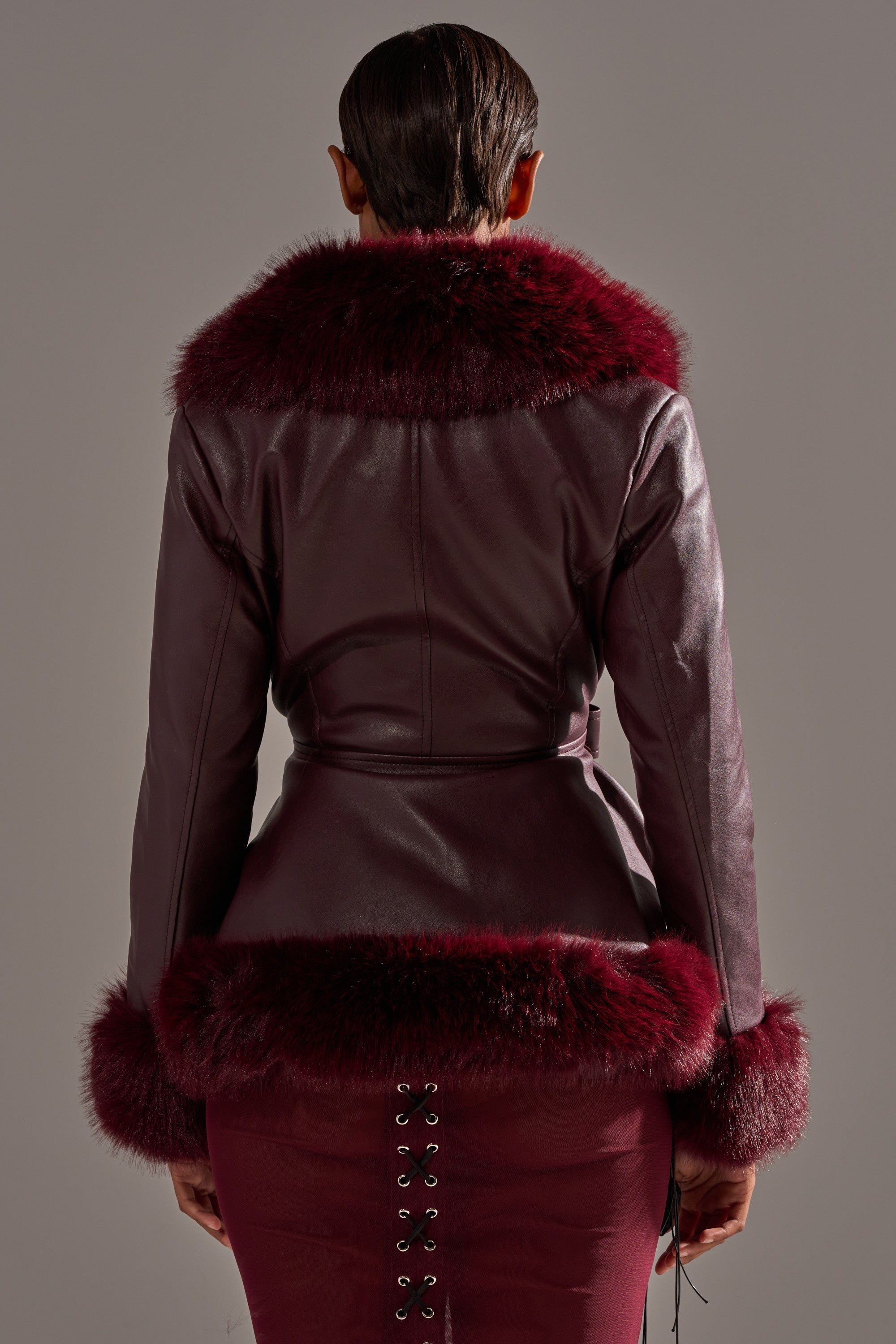 A person is seen from behind wearing the KENZY FUR TRIM MOTO IN BURGUNDY with dark red fur trim on the collar, cuffs, and hem, paired with a matching burgundy skirt featuring lace-up detailing at the back. 5