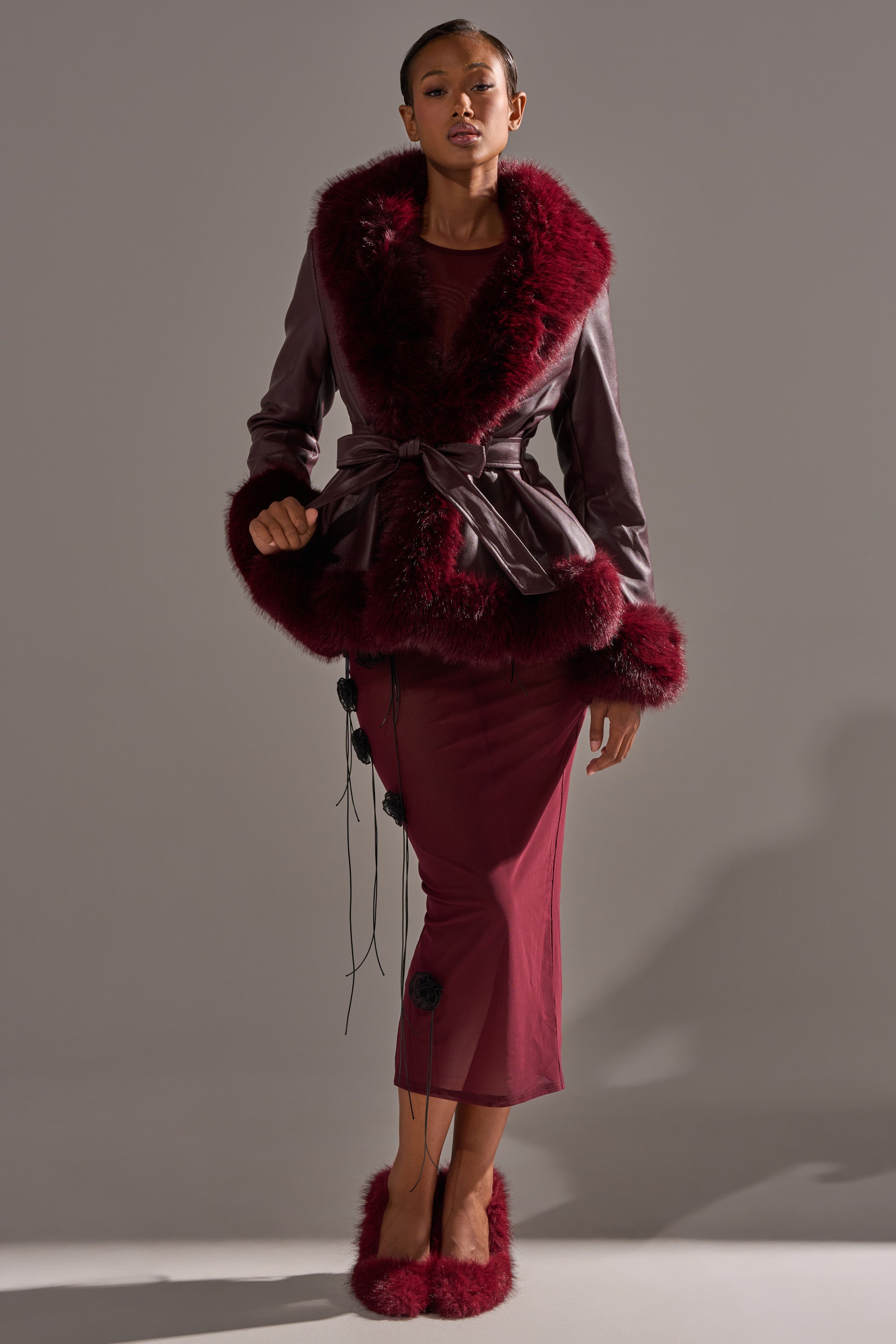 A woman models the KENZY FUR TRIM MOTO IN BURGUNDY—an elegant deep burgundy moto jacket with fur collar and cuffs—over a matching floral dress and fluffy slippers against a plain gray background. 1