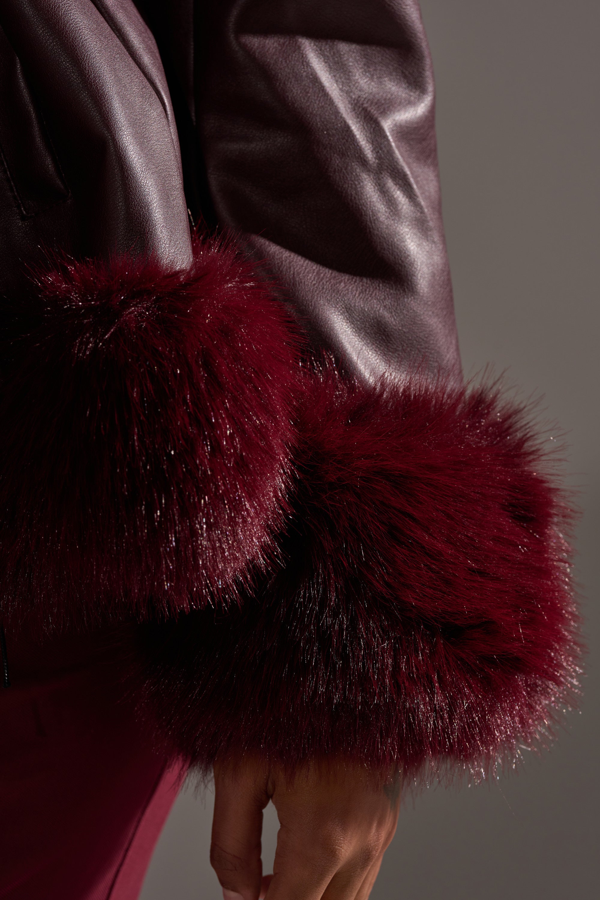 A close-up shows a person’s arm wearing the KENZY FUR TRIM MOTO IN BURGUNDY, featuring dark burgundy faux leather and plush burgundy faux fur cuffs, with their hand relaxed by their side against a neutral background. 8