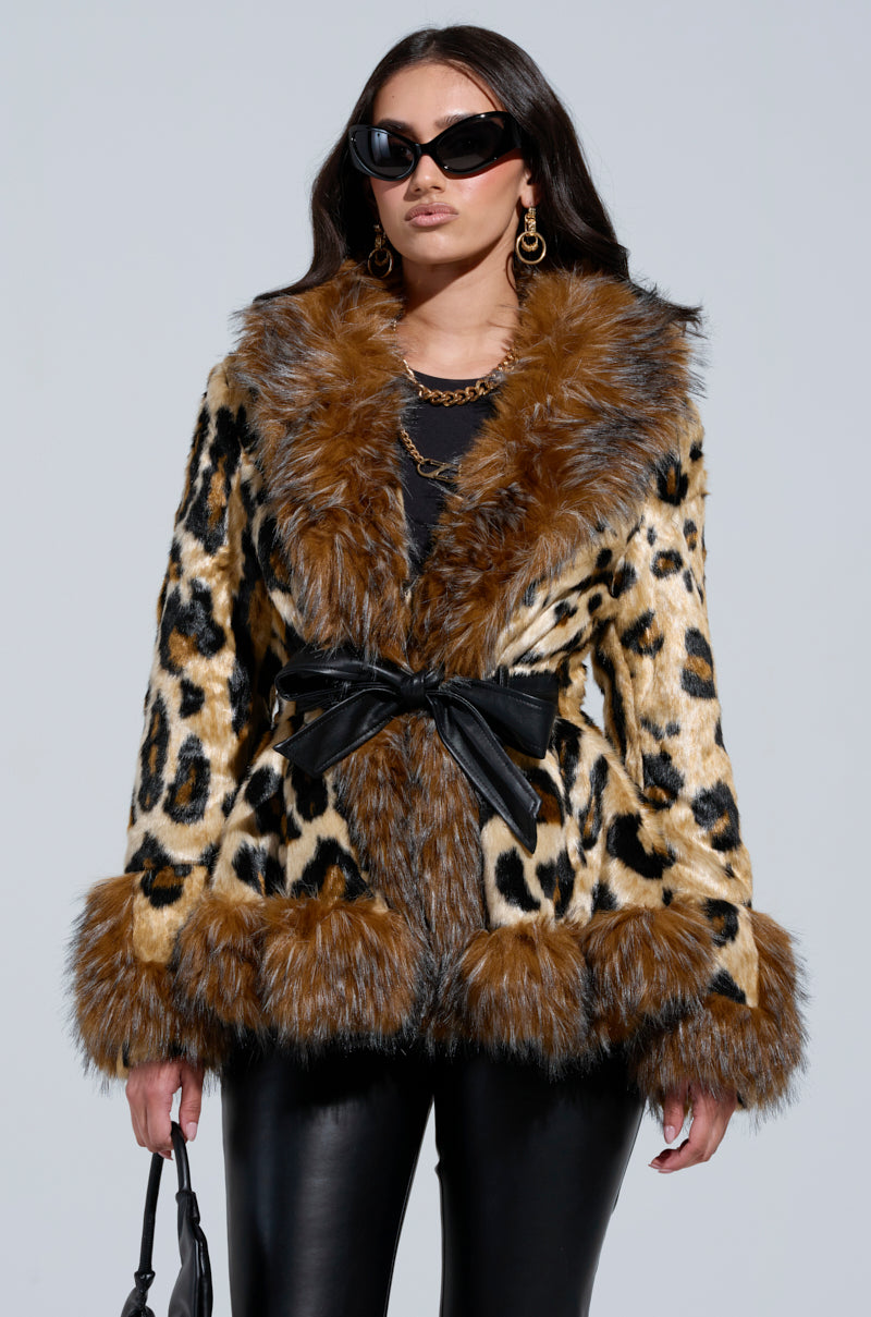 Wearing the KENZY PEPLUM FAUX FUR JACKET, which features an elegant leopard print with faux fur trim and a black belt, a person showcases their fashion-forward style. They complete the look with dark sunglasses and gold earrings, while carrying a black handbag and posing confidently against a plain background.