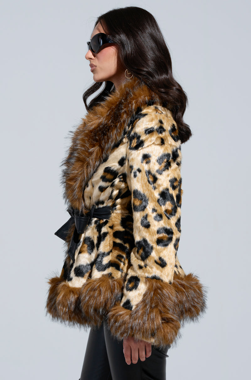 A woman sporting sunglasses and the KENZY PEPLUM FAUX FUR JACKET, featuring a leopard print design with a peplum style and brown fur trim, stands against a plain backdrop. Her long, dark hair flows over her shoulders as she poses in profile, facing left.