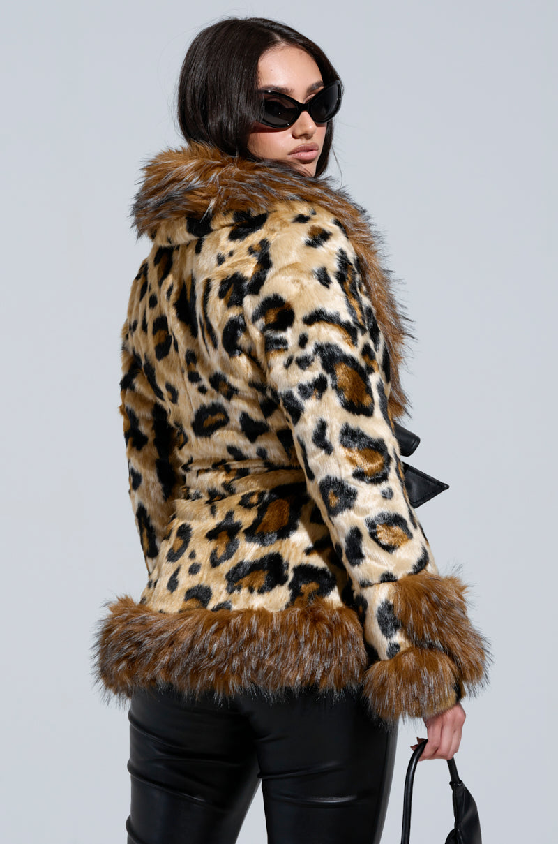 A person wearing dark sunglasses, the KENZY PEPLUM FAUX FUR JACKET with leopard print and brown trim, and black leather pants is facing away while holding a black handbag. The background is a plain light gray.