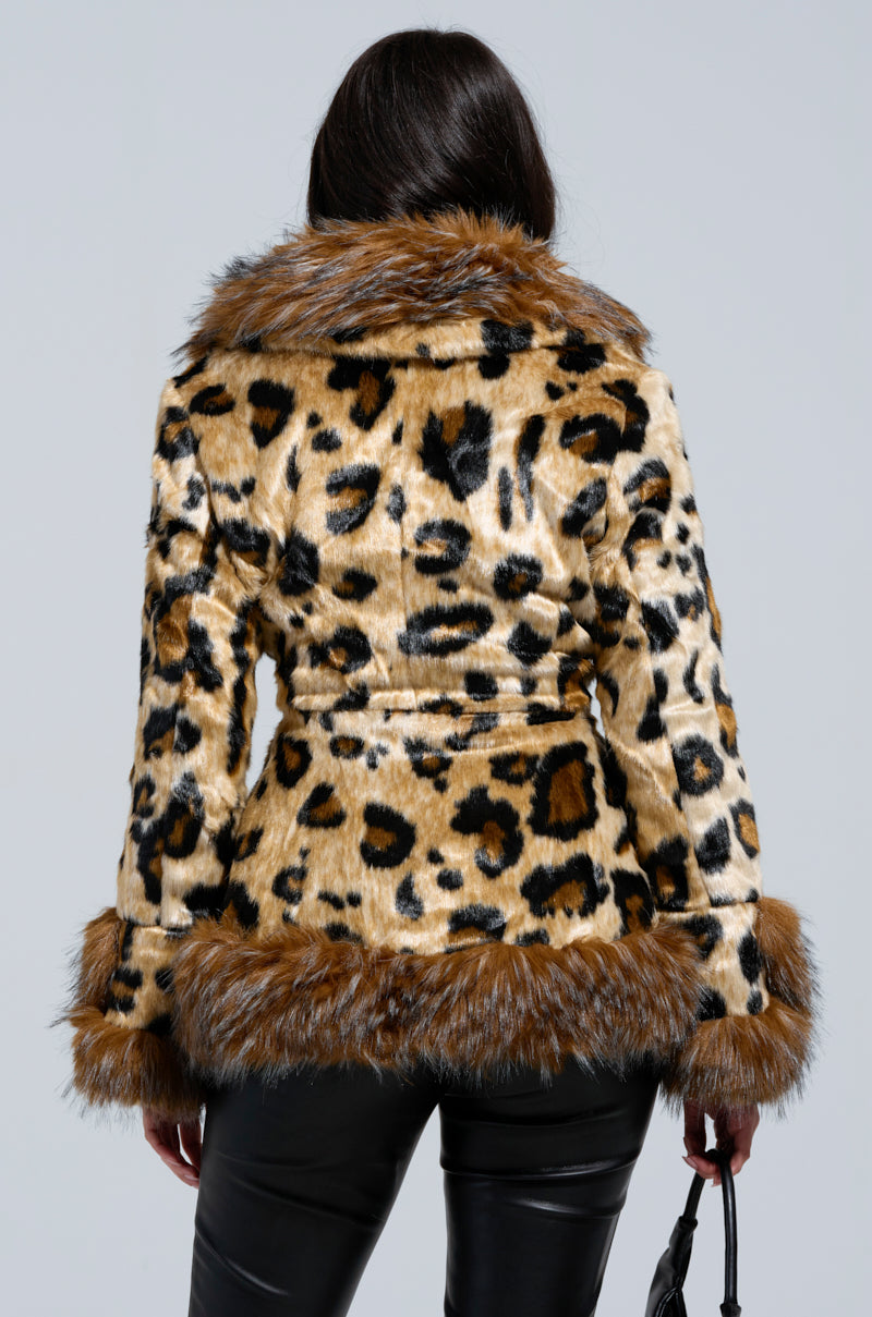 A rear view of an individual wearing the KENZY PEPLUM FAUX FUR JACKET in leopard print, showcasing brown fur trim on the collar and cuffs. The jacket comes with a belt that emphasizes the waist, paired with black pants and accessorized with an elegant black handbag.