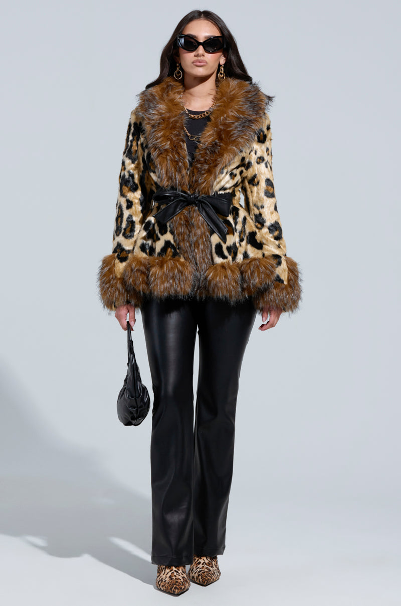 A person wearing the KENZY PEPLUM FAUX FUR JACKET in leopard print, accented with a black belt, complements their look with black leather pants and matching shoes. Their eyes are hidden behind large sunglasses as they hold a small black purse. The jacket's peplum design offers additional flair with fur details on the sleeves and collar.