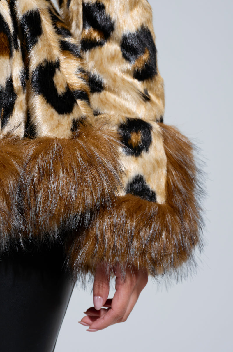 A close-up of a person wearing the KENZY PEPLUM FAUX FUR JACKET, featuring a leopard print design with brown fur trim on the sleeves. The individual stands against a plain background, their hand relaxed by their side.