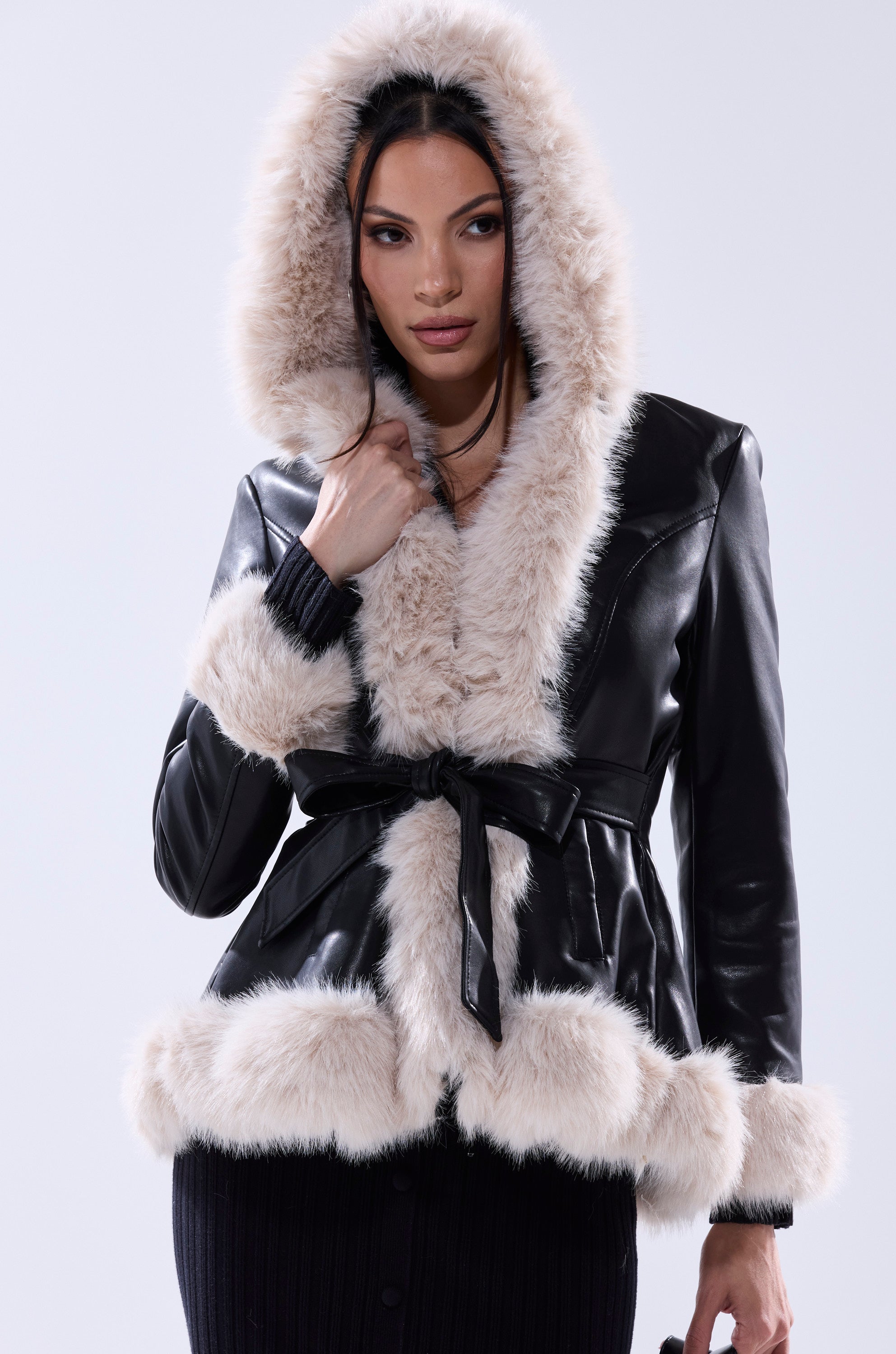 A woman wears the HOODED KENZY FUR TRIM MOTO—a faux leather jacket with light beige fur on the hood, cuffs, and hem, belted at the waist—standing against a plain light background.