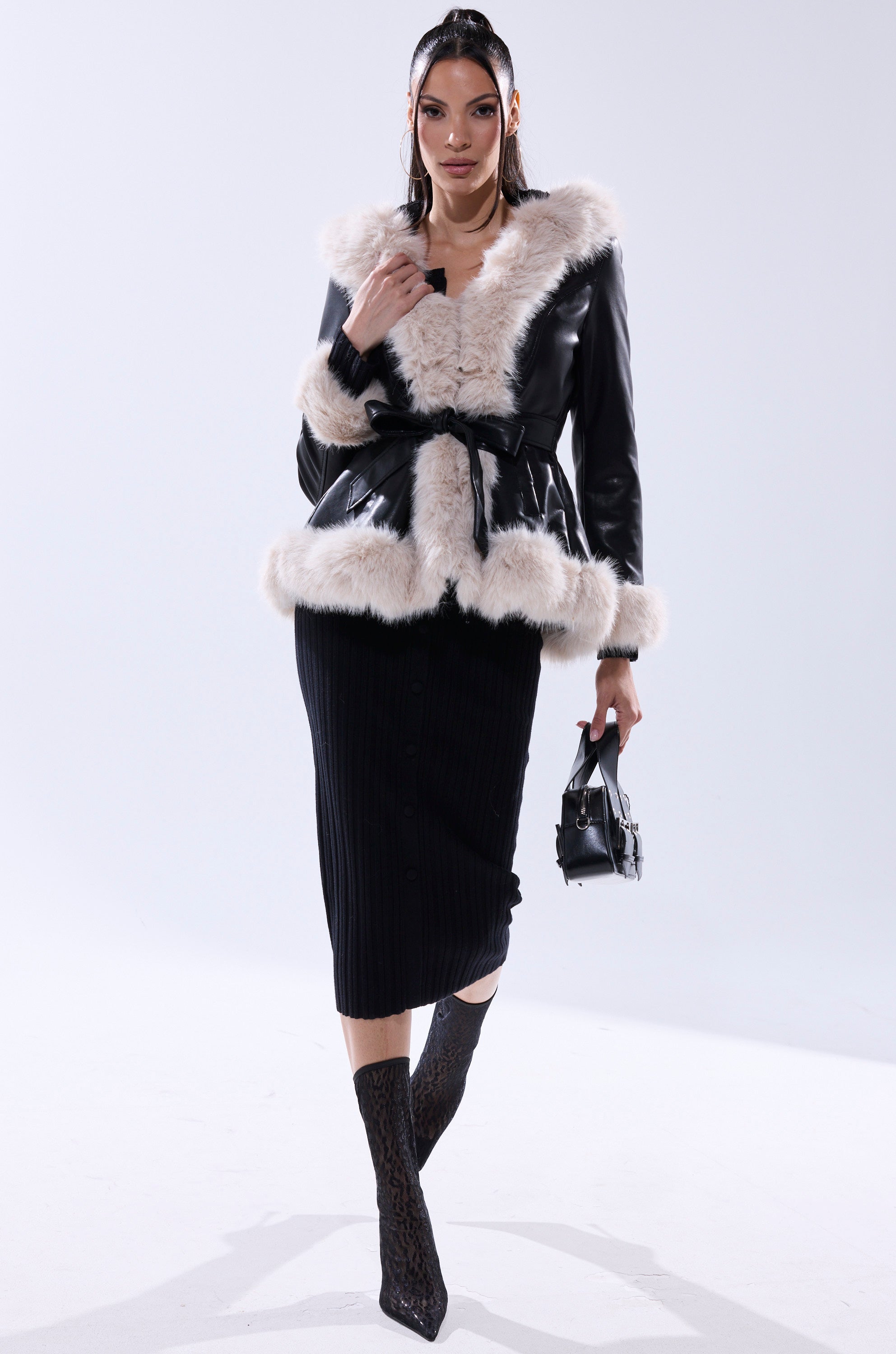 A woman poses in the HOODED KENZY FUR TRIM MOTO jacket with white fur trim, a black ribbed skirt, black lace boots, and a small black handbag against a plain white background.