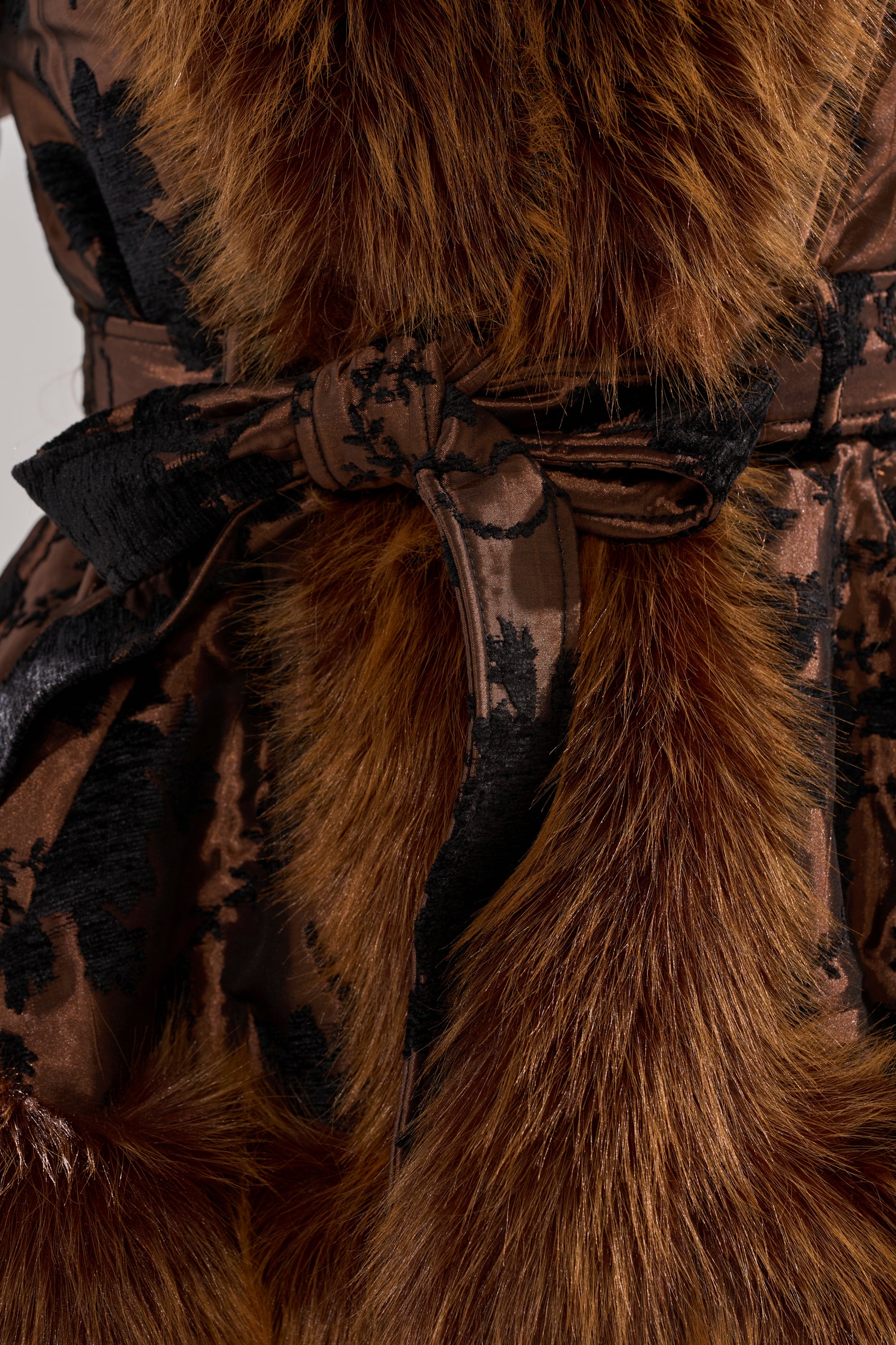 A close-up of the KENZY FUR TRIM MOTO IN JACQUARD shows its soft brown floral jacquard fabric, black floral pattern belt tied in a bow, and luxurious fur trim, highlighting the garment's rich detail and texture.