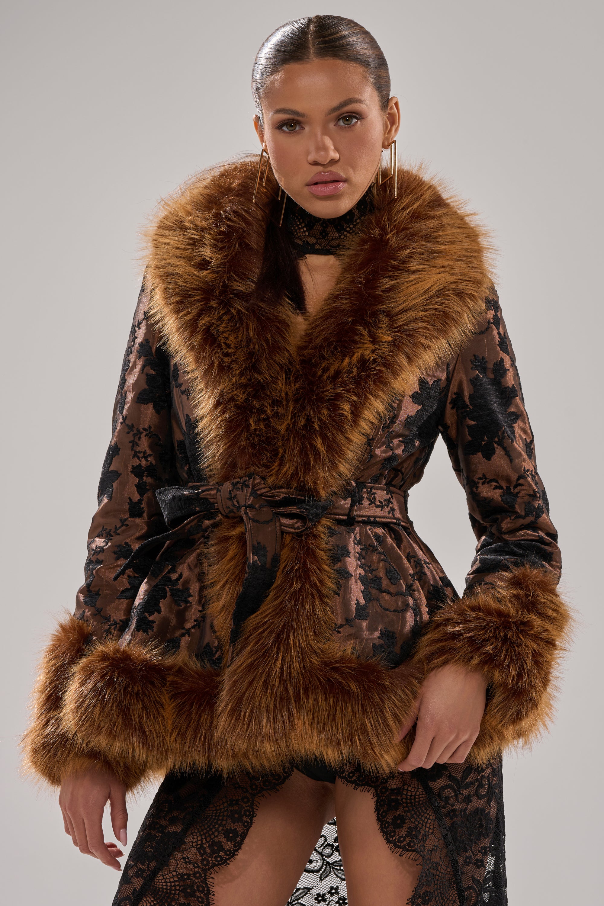 A woman in the KENZY FUR TRIM MOTO IN JACQUARD, featuring a black floral jacquard pattern, brown faux fur collar and cuffs, tie belt waist, and a lacy black skirt, stands against a plain gray background.
