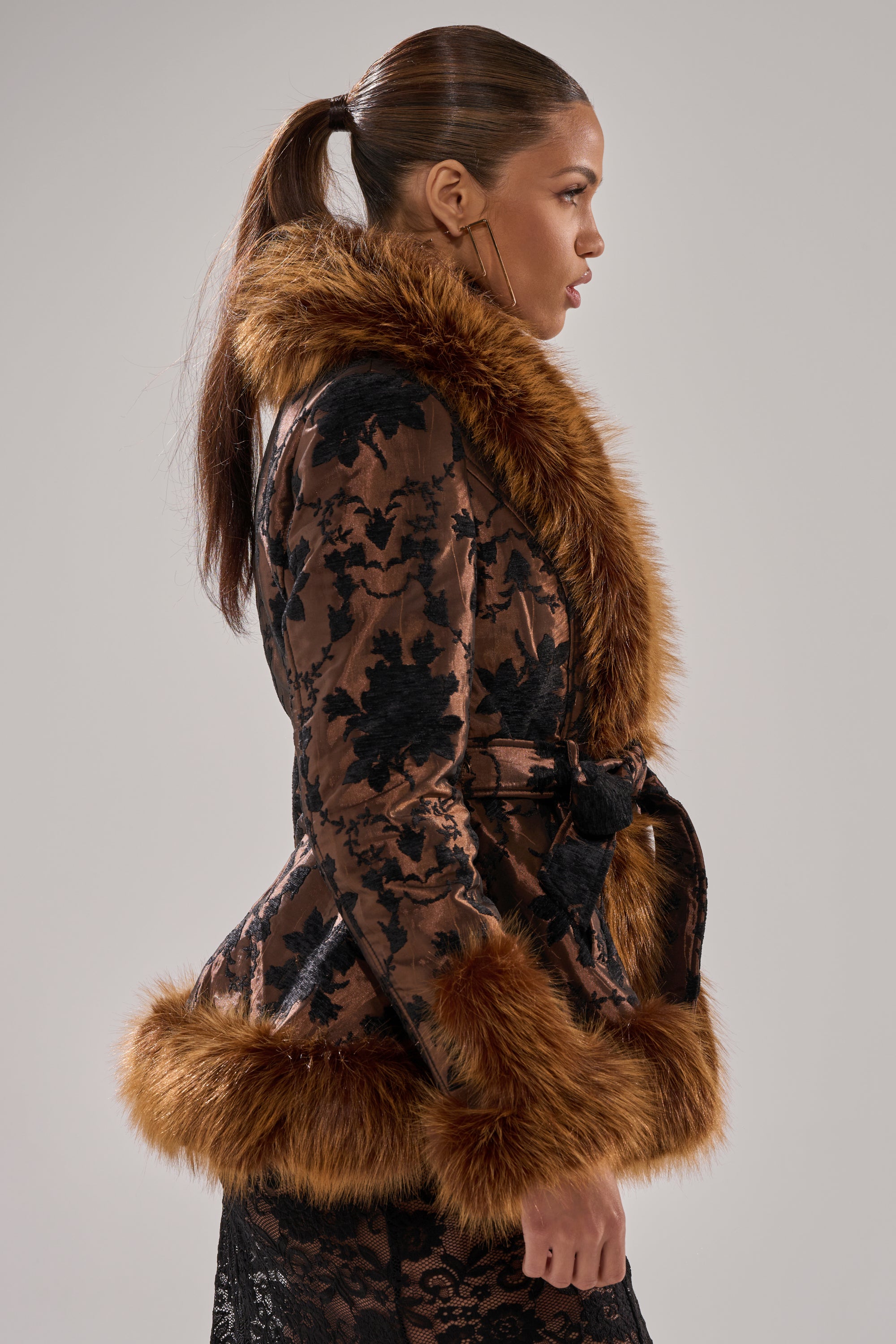 A woman in profile, hair in a ponytail, stands against a plain background wearing the KENZY FUR TRIM MOTO IN JACQUARD—a floral jacquard coat featuring faux fur at the collar, cuffs, and hem.