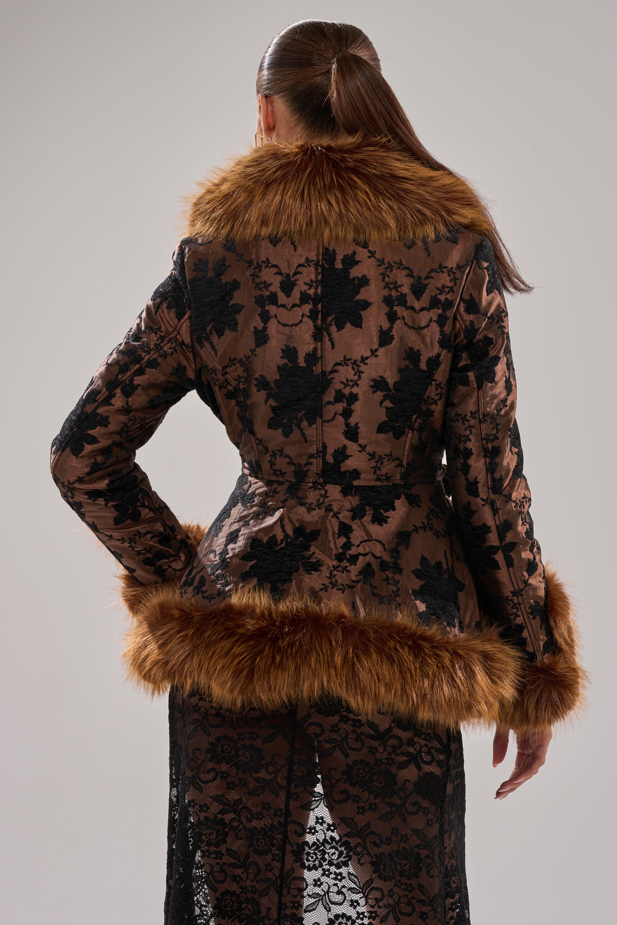 A woman with her hair in a sleek ponytail stands with her back to the camera, wearing the KENZY FUR TRIM MOTO IN JACQUARD, featuring a floral jacquard design and faux fur collar, cuffs, and hem.