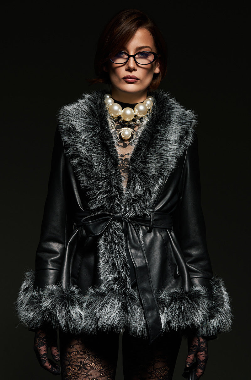 A person wearing the "NEW KENZY FUR TRIM MOTO" black faux leather coat with gray sherpa lining stands against a dark background. They have short brown hair, glasses, and a large pearl necklace. The coat is belted at the waist, and they are wearing lace stockings.