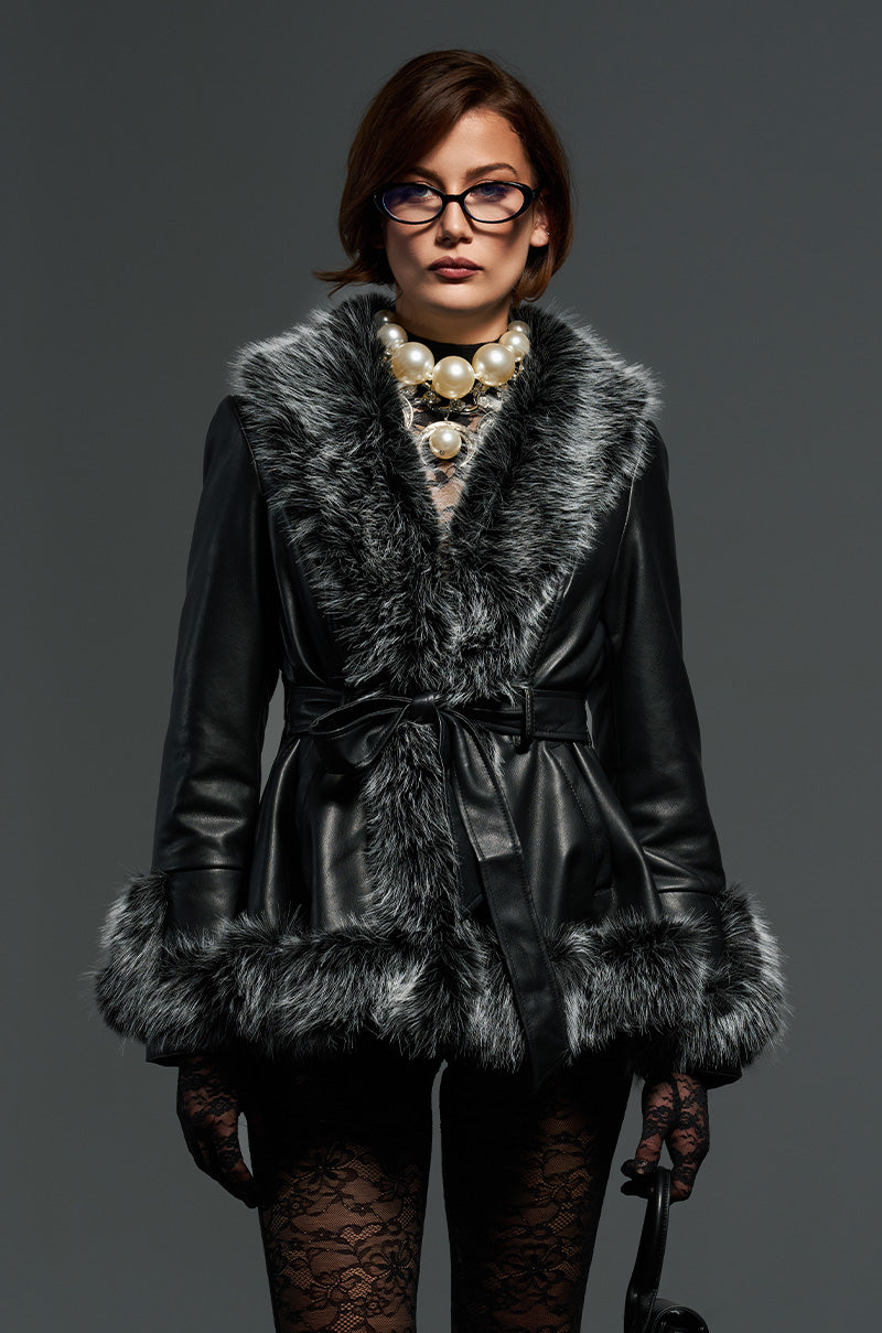 A woman confidently poses in the NEW KENZY FUR TRIM MOTO jacket. She has short hair, wears glasses, statement pearl jewelry, and lace gloves while standing against a gray background.