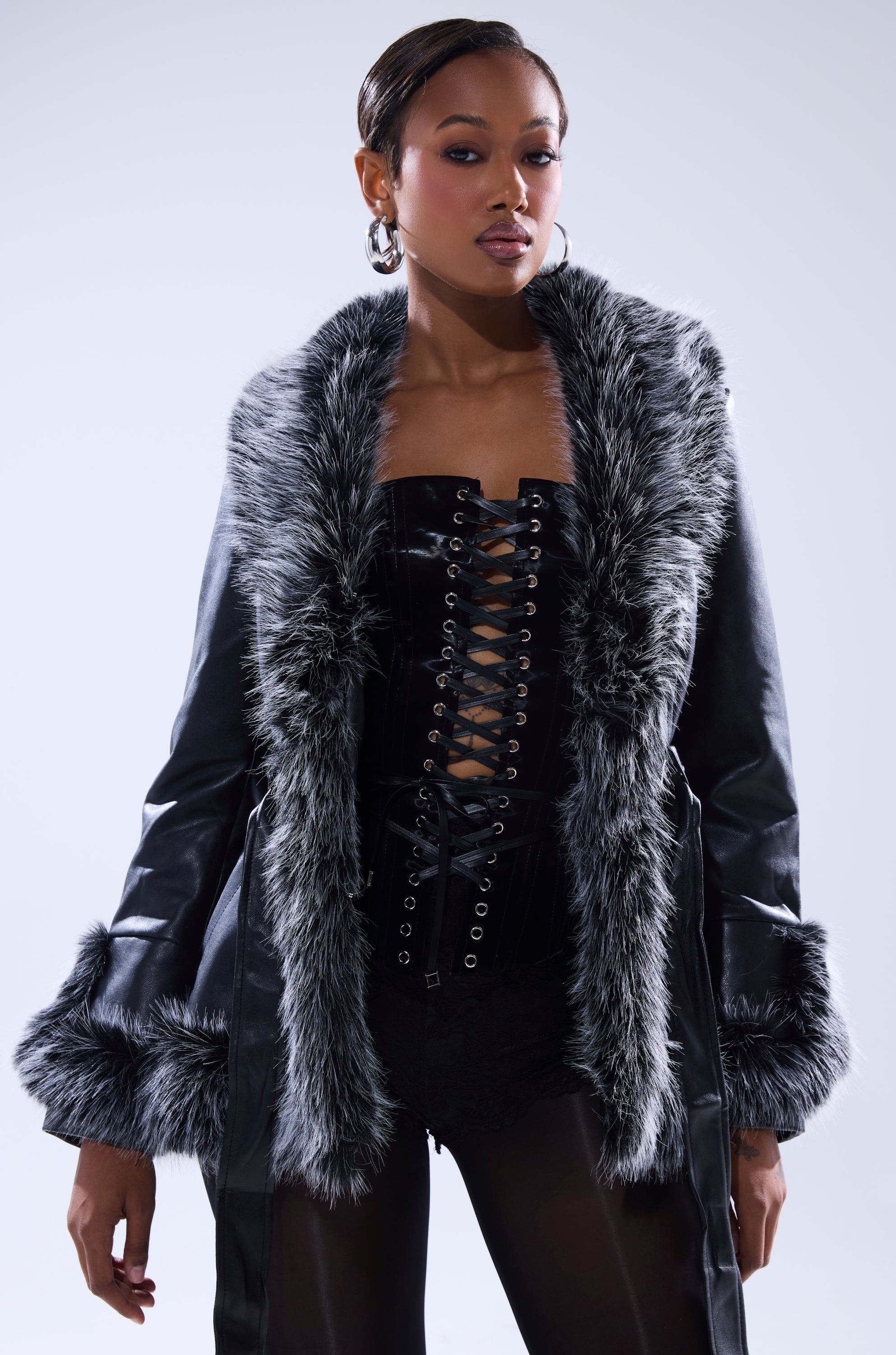 A woman stands confidently against a light background in the NEW KENZY FUR TRIM MOTO, a black faux leather moto jacket with grey fur trim over a black lace-up corset top, exuding edgy sophistication.