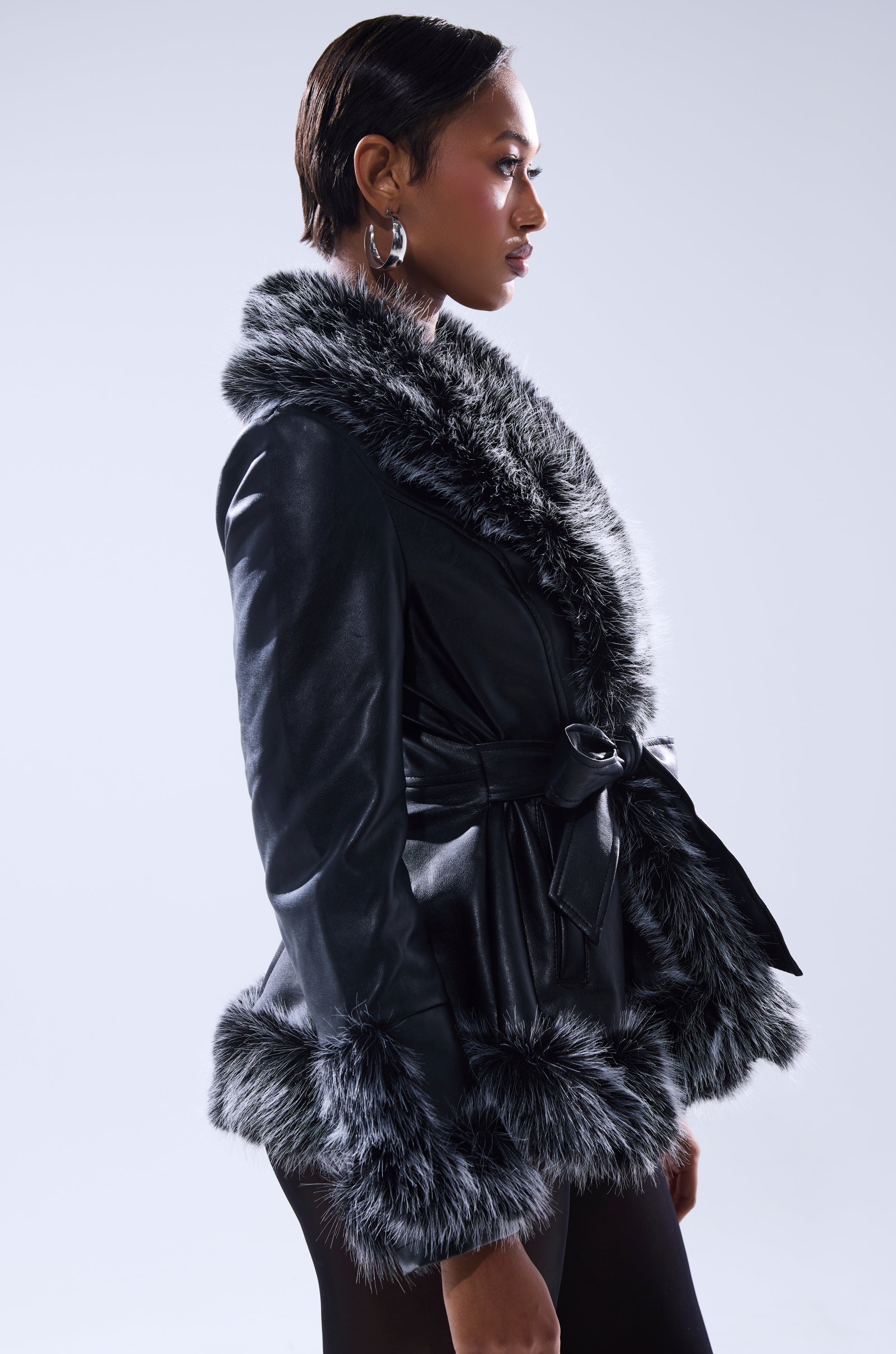 A woman in profile models the NEW KENZY FUR TRIM MOTO, a black faux leather jacket with thick gray fur trim and a belted waist. She has short hair, wears hoop earrings, and stands against a light background.