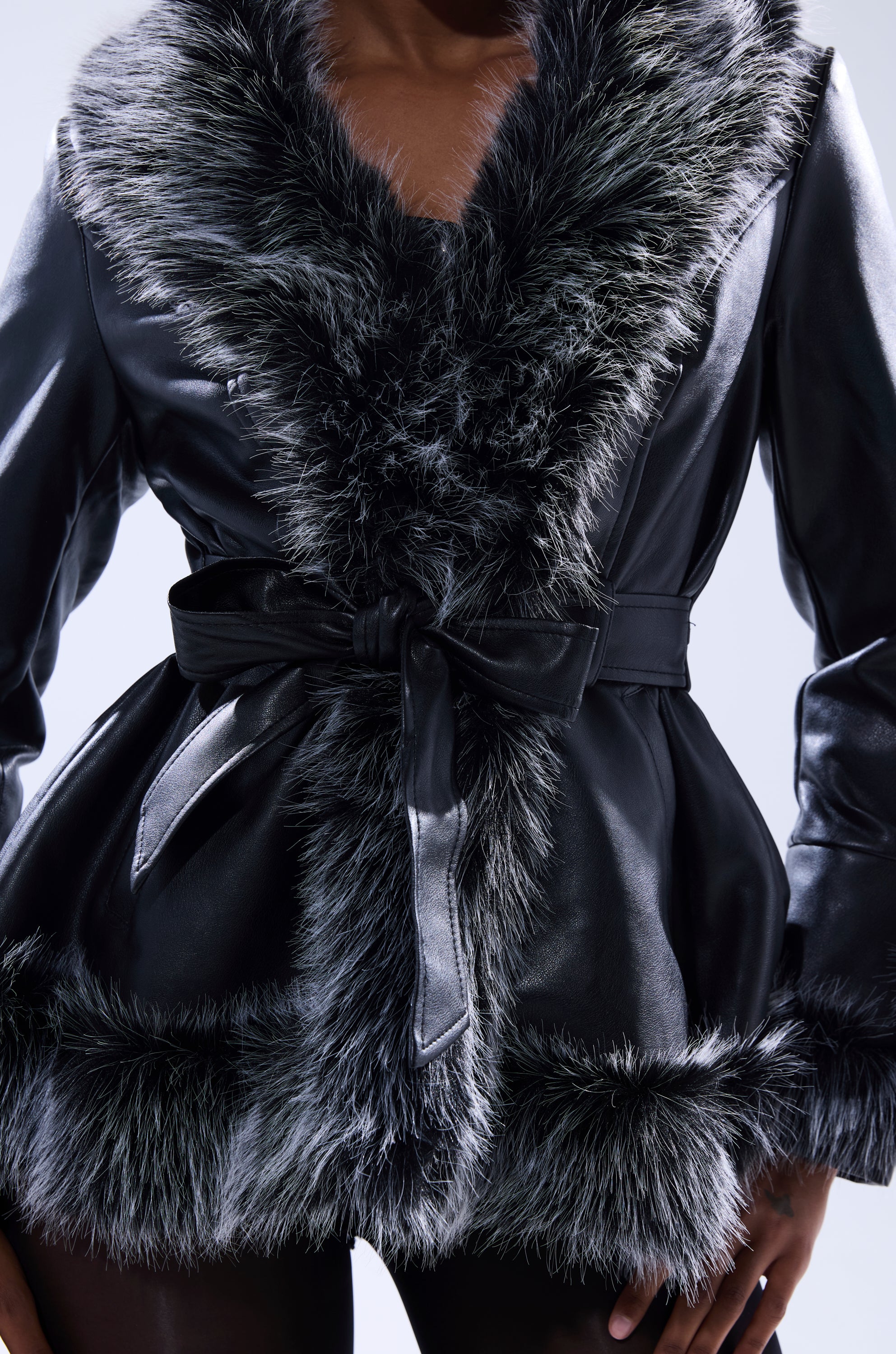 A person wears the NEW KENZY FUR TRIM MOTO, a black faux leather jacket with wide fur trim at the collar and hem, tied at the waist with a matching belt. The cropped longline style is shown from shoulders to mid-thigh.
