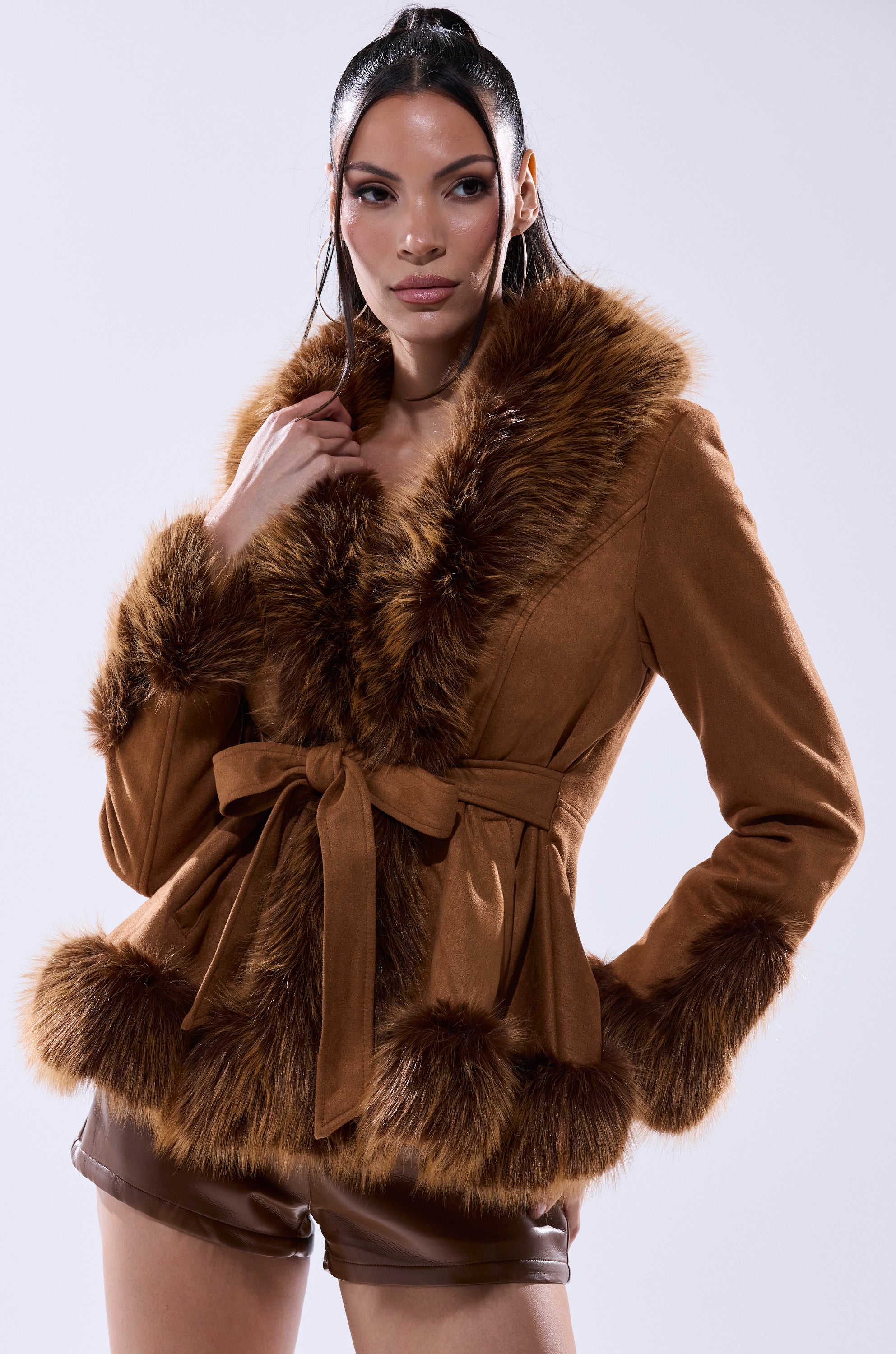 A woman models the SUEDE KENZY FUR TRIM MOTO jacket with brown faux fur trim, a matching belt, and brown shorts as she poses confidently against a plain light background.