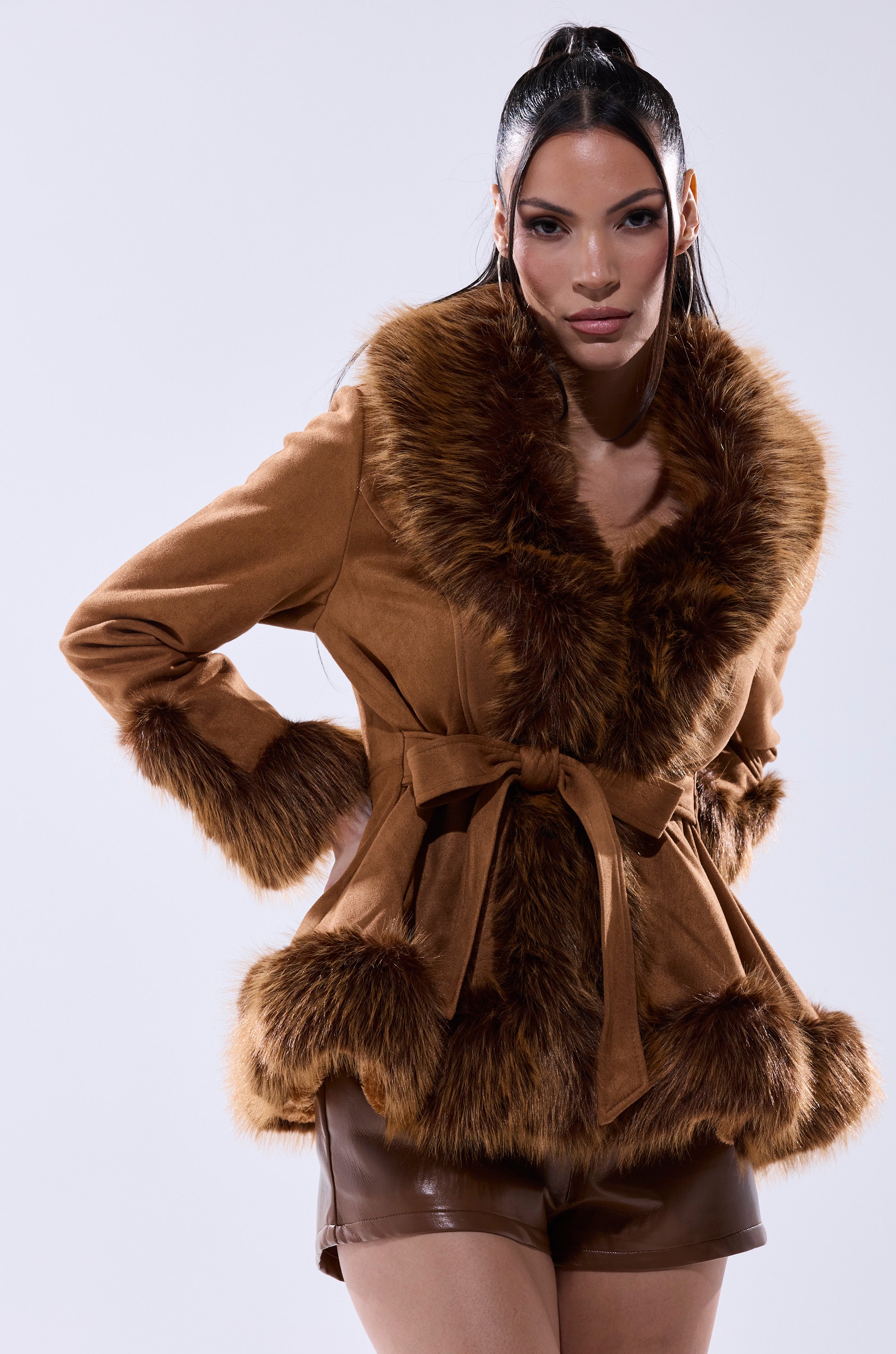 A woman wears the SUEDE KENZY FUR TRIM MOTO jacket with a waist tie and brown shorts, posing confidently against a plain light background.