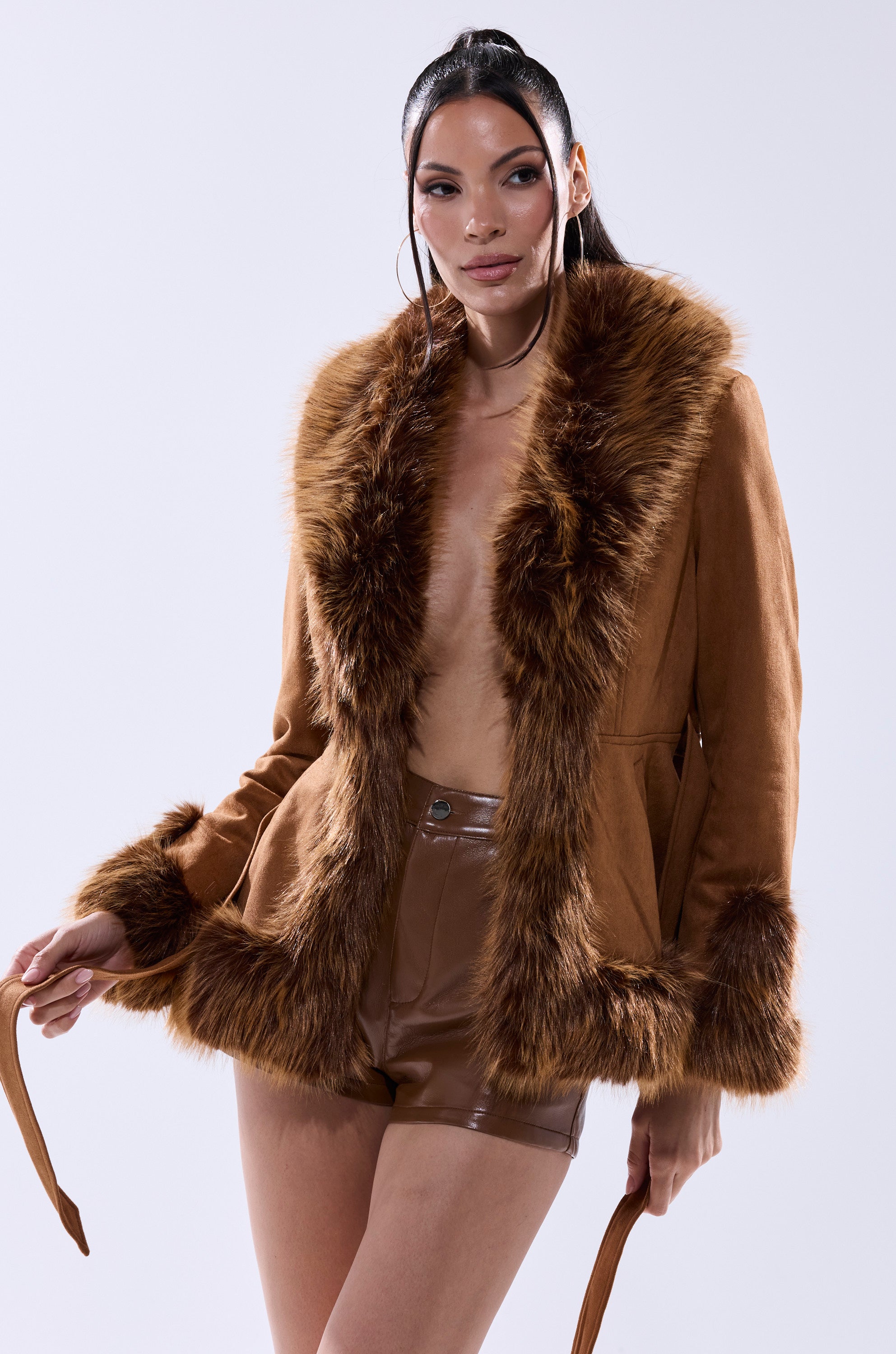 A woman with long dark hair confidently poses in the SUEDE KENZY FUR TRIM MOTO jacket and brown leather shorts against a plain light background.