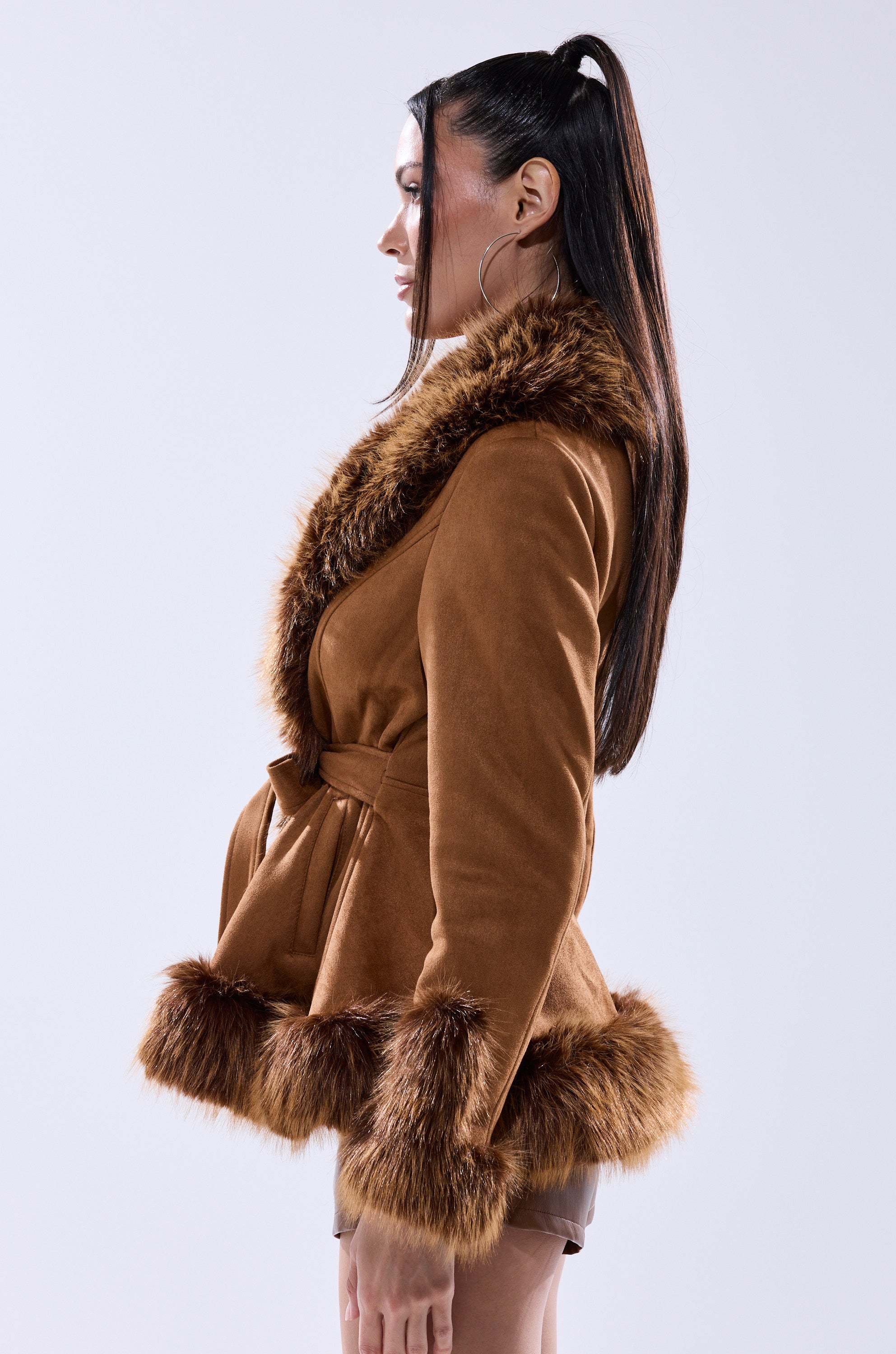 A woman with long dark hair in a high ponytail wears the SUEDE KENZY FUR TRIM MOTO jacket, featuring vintage-inspired faux fur trim on the collar, cuffs, and hem, as she stands in profile against a plain light background.