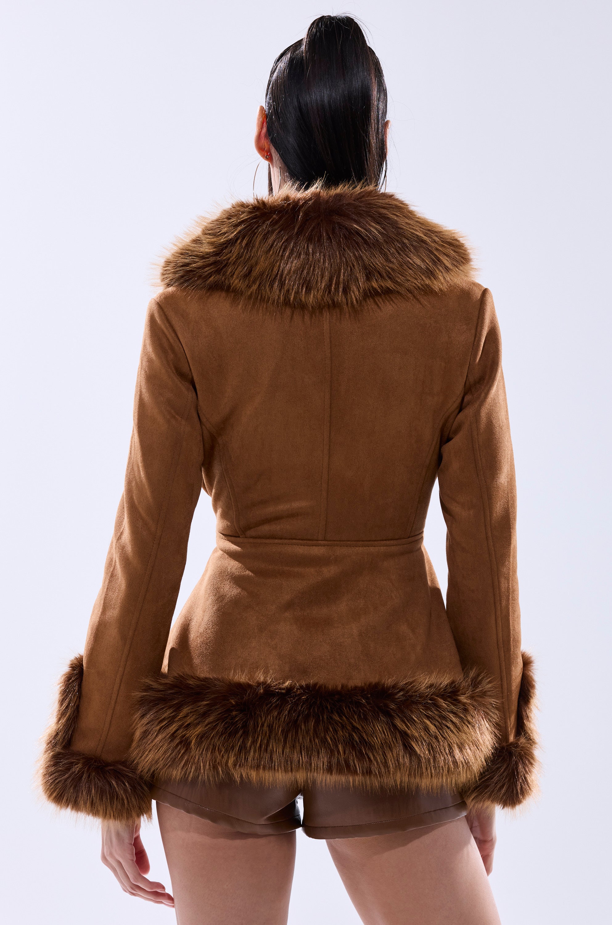 A person stands facing away, wearing the SUEDE KENZY FUR TRIM MOTO jacket—brown faux suede with fur trim on the collar, cuffs, and hem—paired with beige shorts against a plain, light-colored background.
