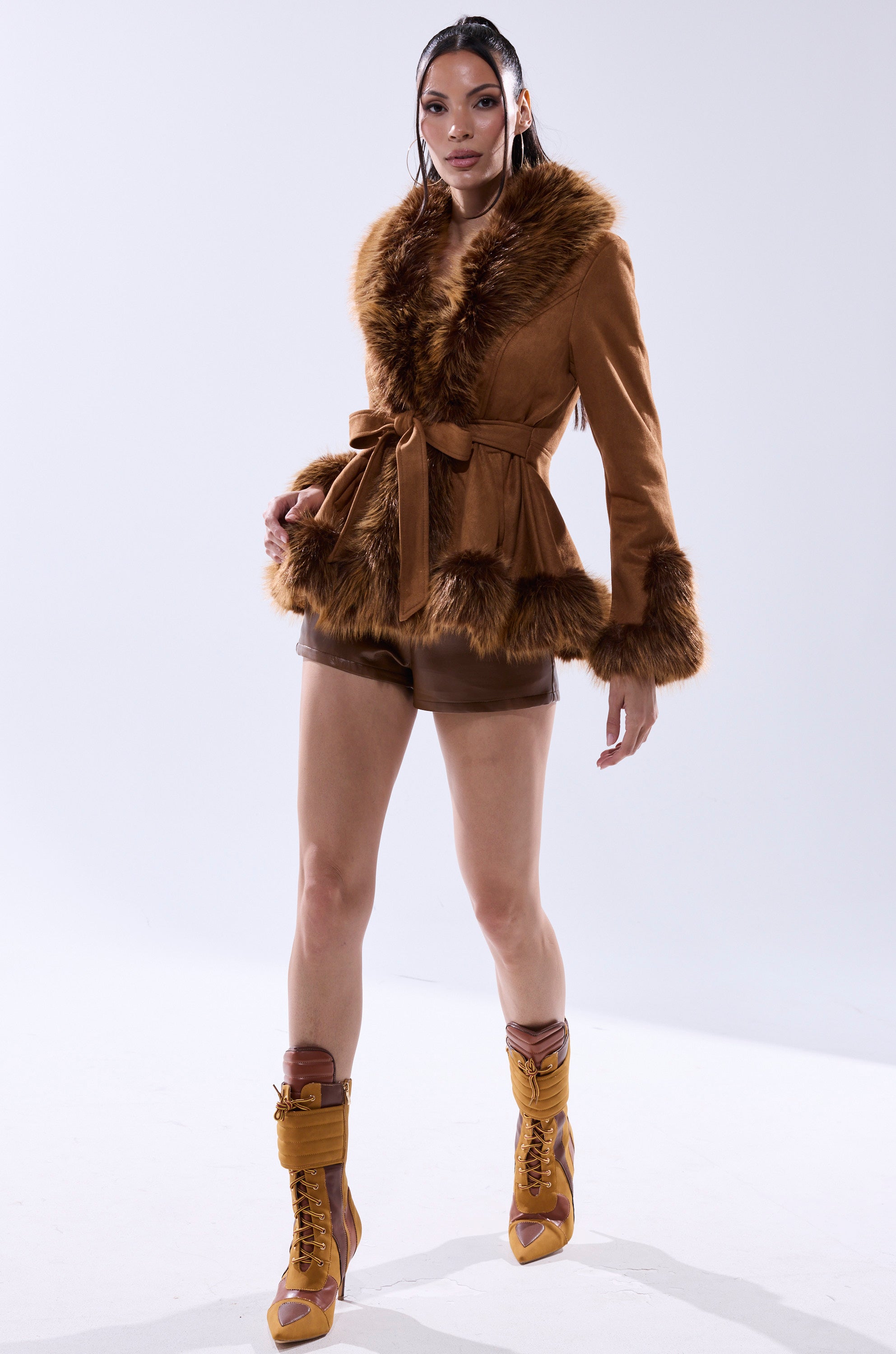 A woman poses against a white background wearing the SUEDE KENZY FUR TRIM MOTO jacket with a tied waist, styled with brown shorts and tan lace-up heeled boots.