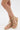 A person shows off the AKIRA KEONA BEIGE ROPE FLAT SANDAL, featuring a strappy criss-cross design with multiple knots, while standing on a white surface. Only their lower legs and feet are visible.