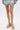 A person stands on a white surface, showing only their legs and feet. They are wearing the AKIRA KEONA BEIGE ROPE FLAT SANDAL, featuring strappy criss-cross design with thick, knotted beige rope straps.
