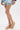 Only the lower half of a person is visible as they stand on a white background, wearing a colorful dress paired with AKIRA KEONA BEIGE ROPE FLAT SANDALS, featuring strappy criss-cross rope and knotted ankle details.