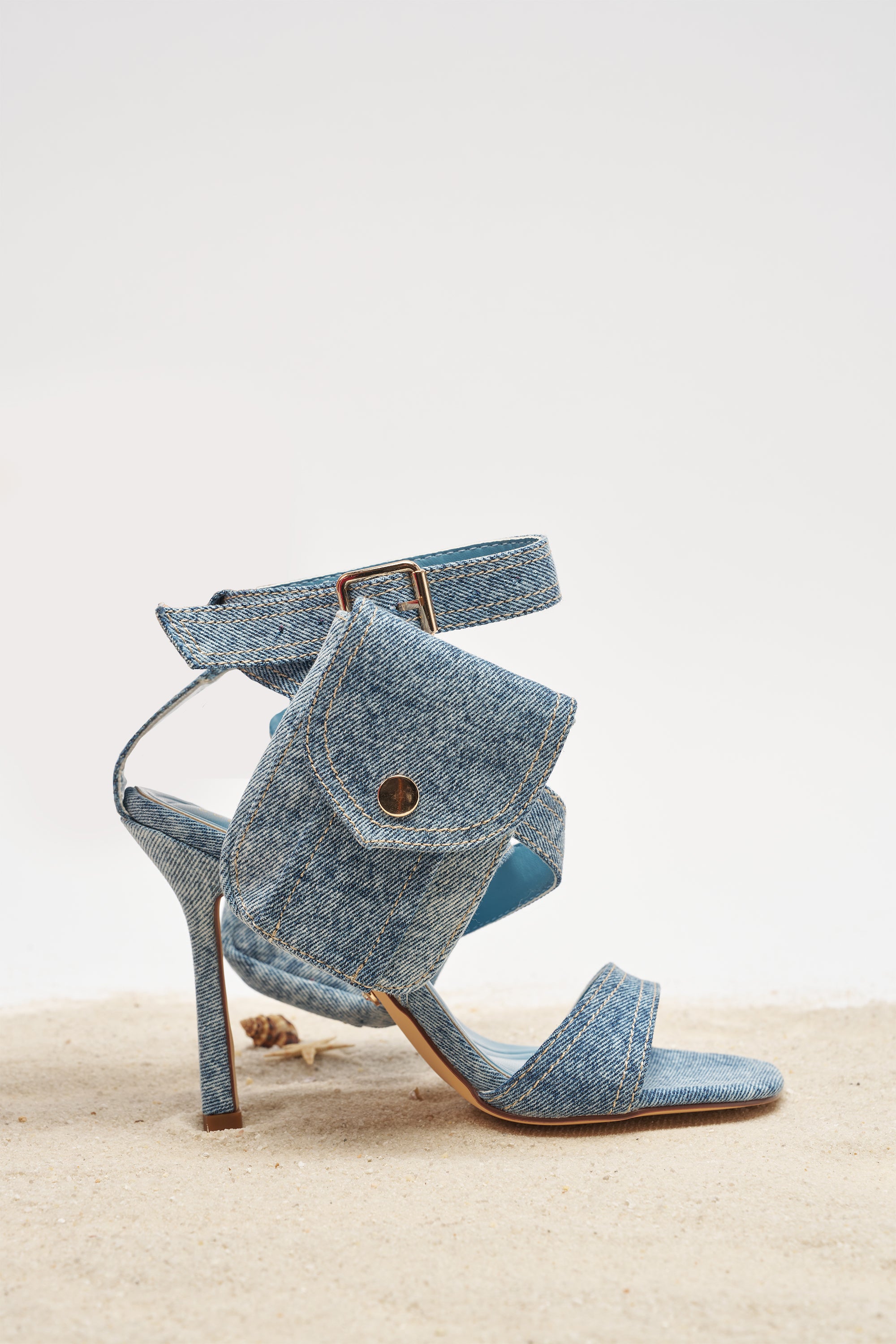 The AKIRA GOLD KERSHAW DENIM POCKET STILETTO STRAP SANDAL is a blue denim sandal with an ankle strap and small side cargo pocket, shown on sandy ground with a plain background. 1