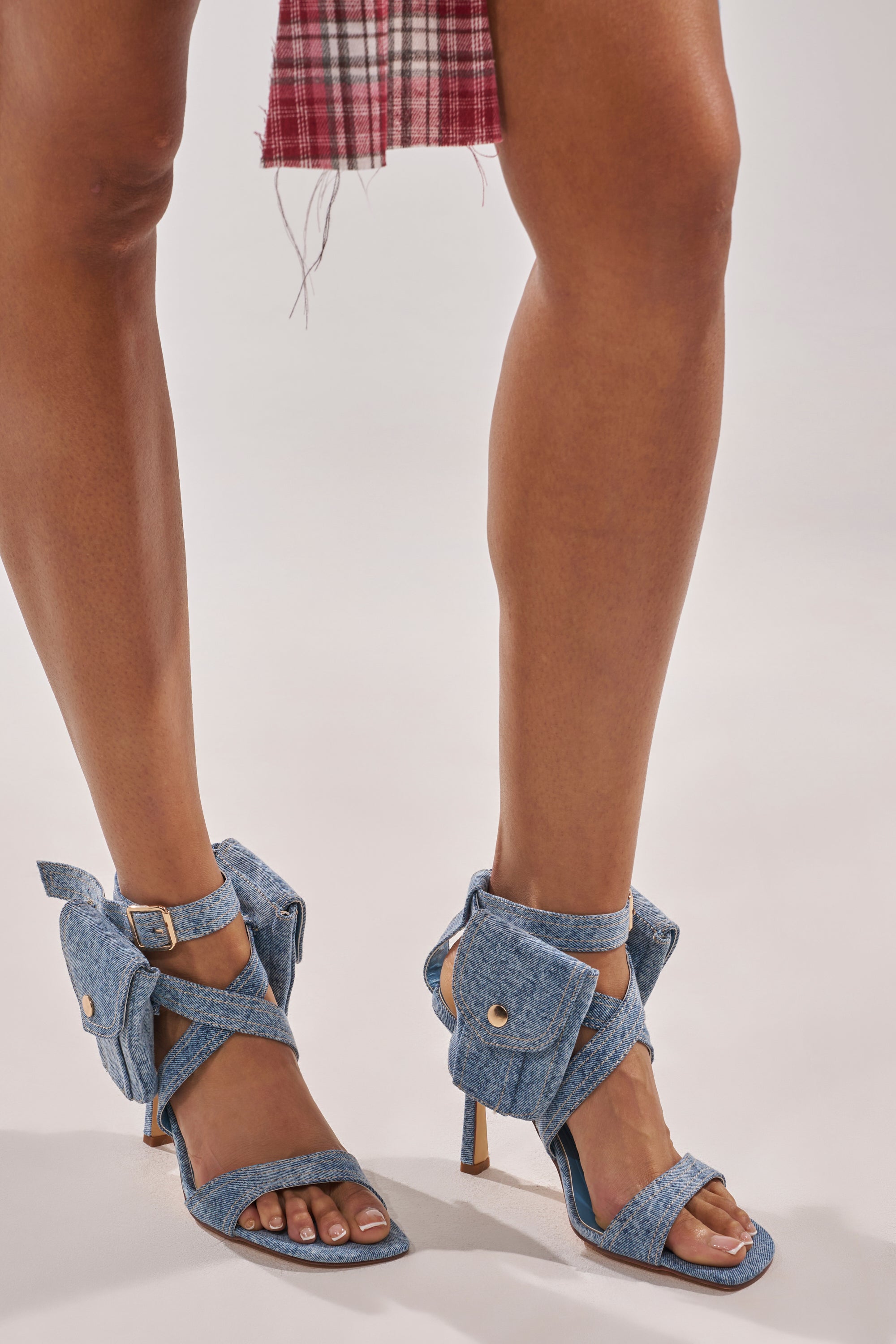 Wearing the AKIRA GOLD KERSHAW DENIM POCKET STILETTO STRAP SANDAL with criss-cross ankle straps and cargo pocket details, paired with a red plaid skirt, she shows off unique textured blue denim heels. 3