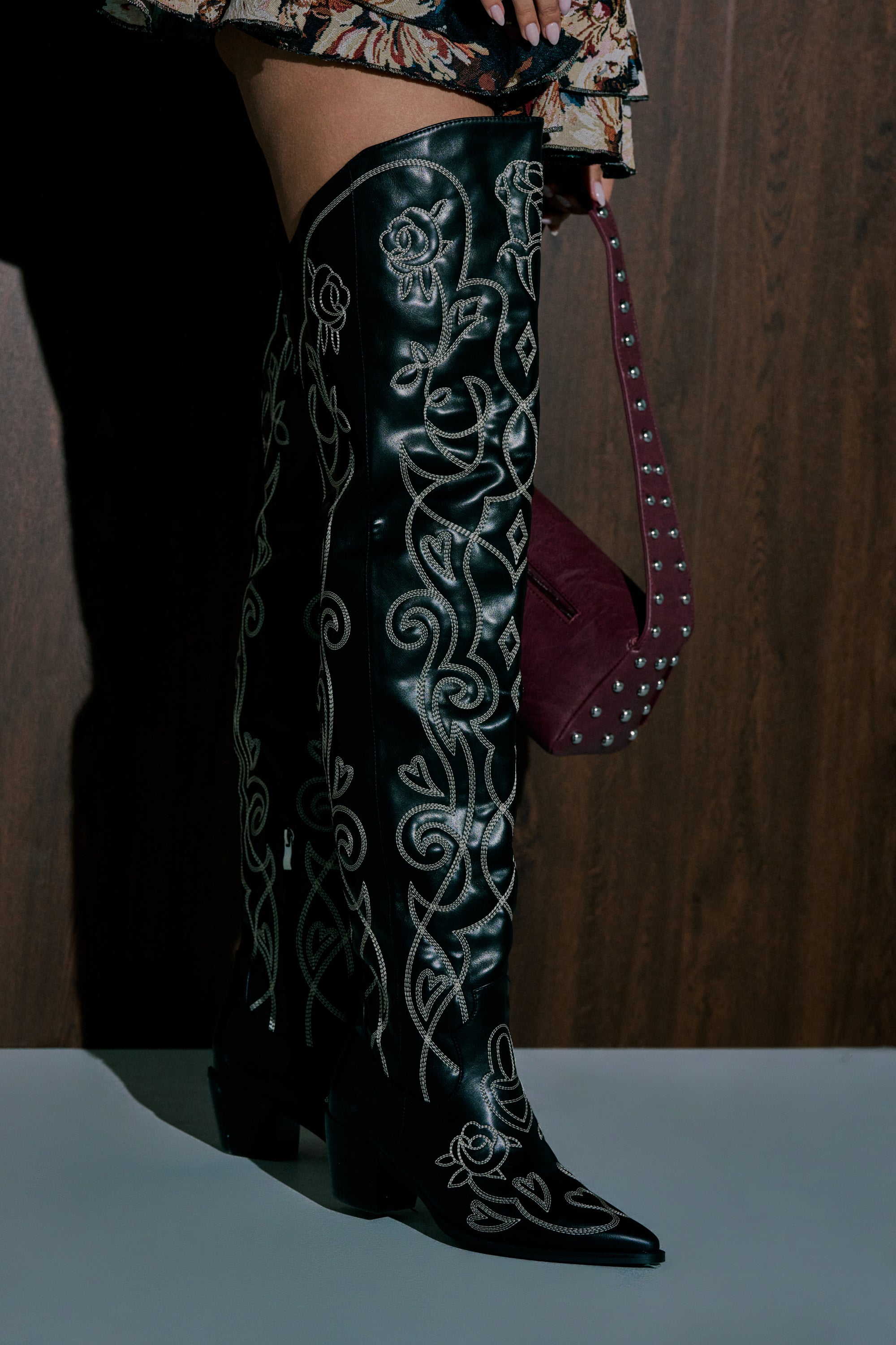 A person stands on a white floor wearing the AZALEA WANG KESS BLACK WESTERN THIGH HIGH CHUNKY BOOT with intricate white embroidery, paired with a floral skirt and holding a burgundy studded handbag. 1