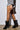 A person stands sideways wearing a houndstooth jacket and pleated skirt, highlighting their legs in sheer black AZALEA WANG ELLIS BLACK BOOTIE knee-high boots with pointed toes and stiletto heels. The backdrop is a plain, light-colored wall and floor.