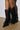 Someone is wearing the AZALEA WANG ELLIS BLACK BOOTIE, knee-high with black mesh, pointed toes, and intricate lace-up details. The striking stiletto heels rest on a light brown floor against a pristine white wall.
