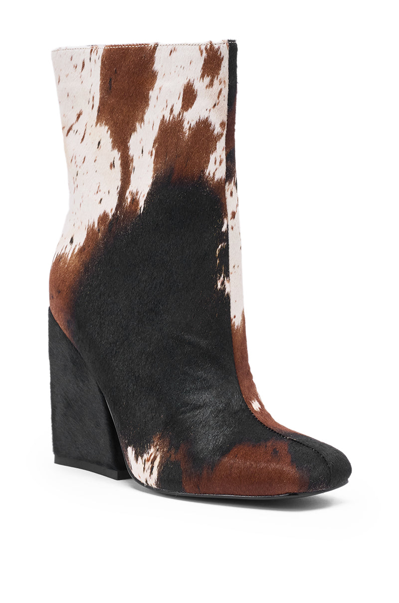The AZALEA WANG KIENNA COW CHUNKY BOOTIE features a pointed toe, thick angular heel, and a black, brown, and white cowhide pattern in soft calf hair, blending unique style with a cozy, chunky-heeled design.